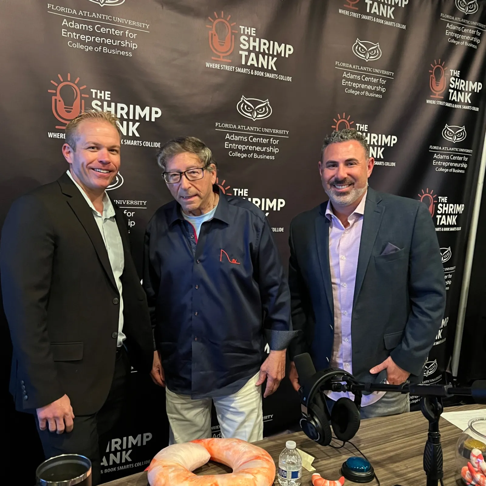Three men posing in front of The Shrimp Tank podcast backdrop with shrimp-themed props and microphones on a table