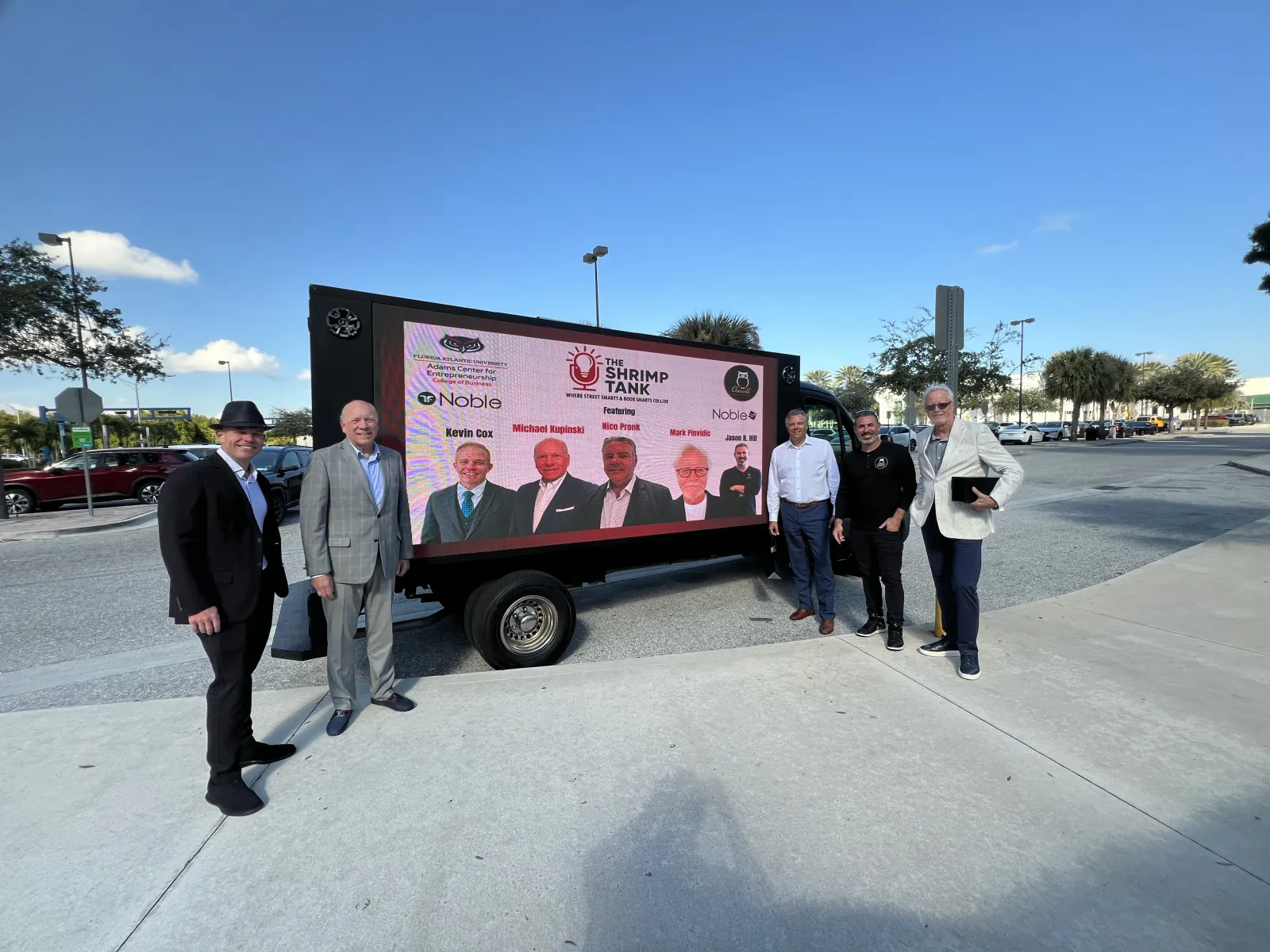 Six men in business attire stand outdoors beside a large digital screen advertising The Shrimp Tank event.