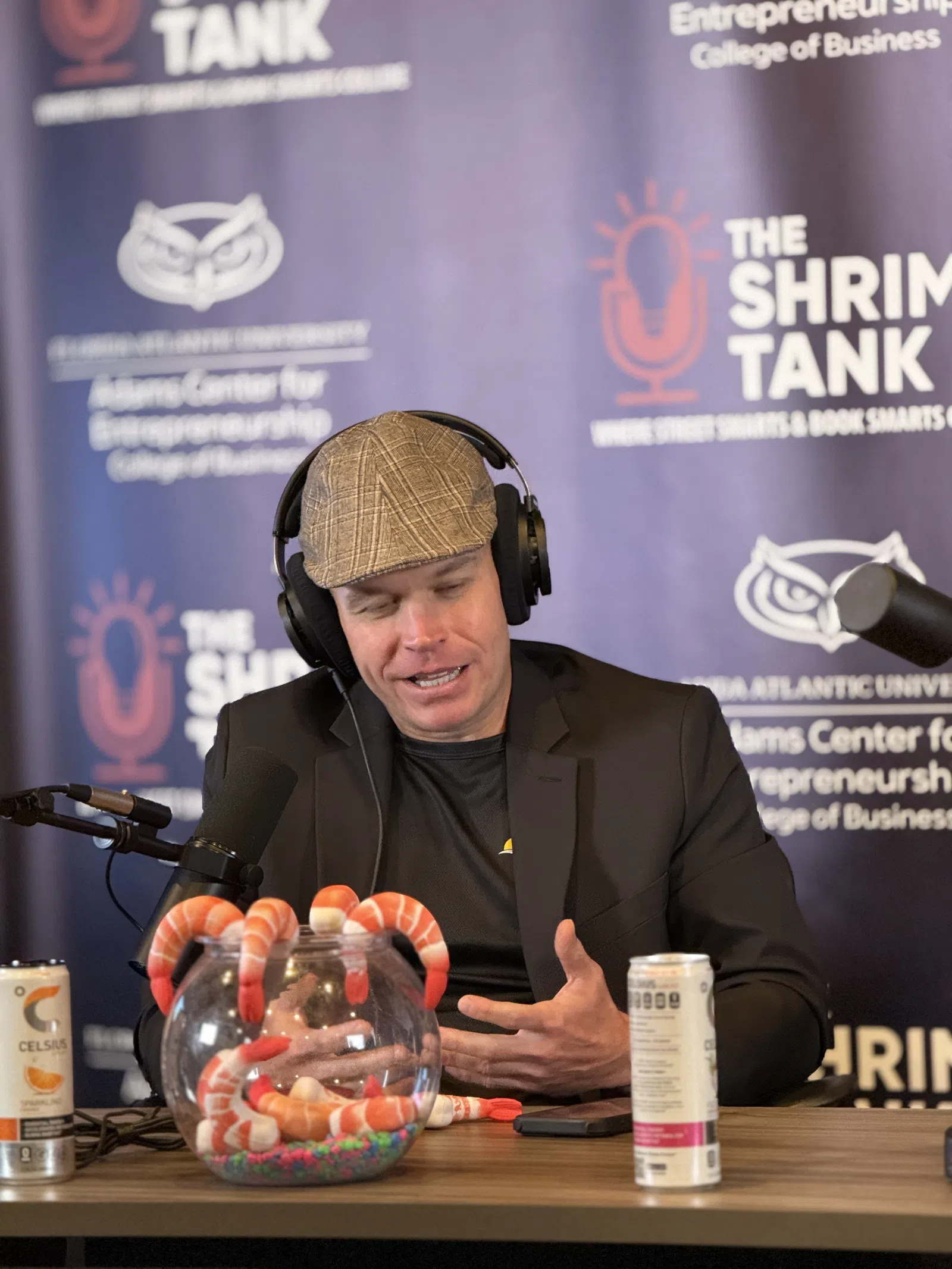 Man wearing headphones and a cap speaking into a microphone at a podcast setup with shrimp-themed decorations and energy drinks.