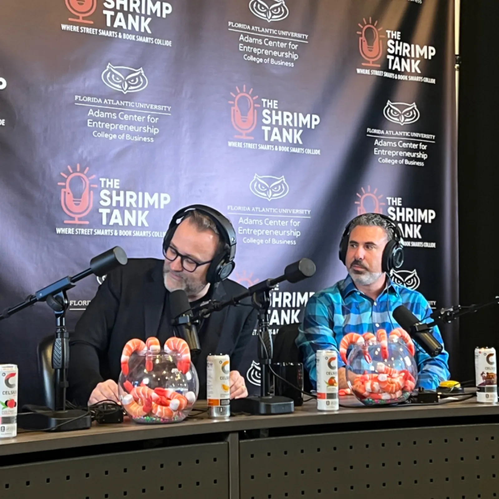 Two men with headphones recording The Shrimp Tank podcast with branded backdrop and shrimp bowls on the desk