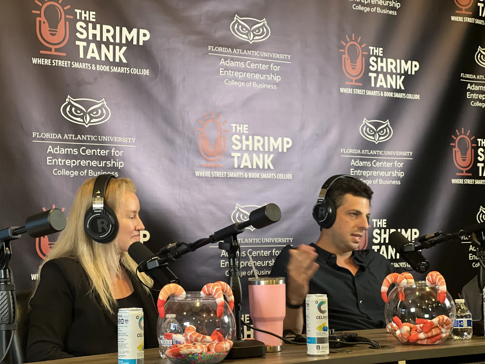 Two podcasters with headphones talk into microphones in front of a backdrop for The Shrimp Tank podcast at Florida Atlantic University.