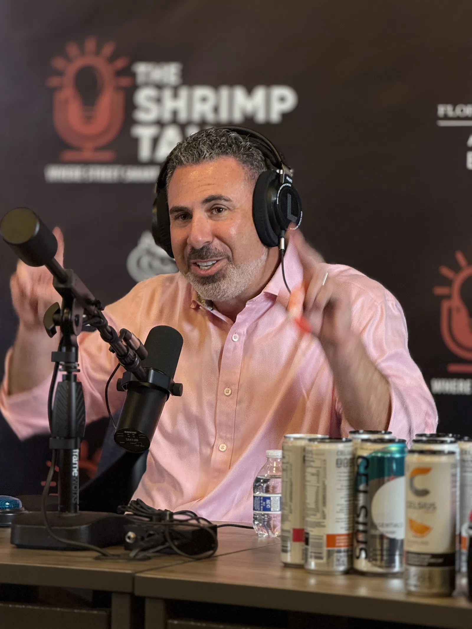 Podcast host wearing headphones speaking into a microphone with energy drinks and water on the table in front.