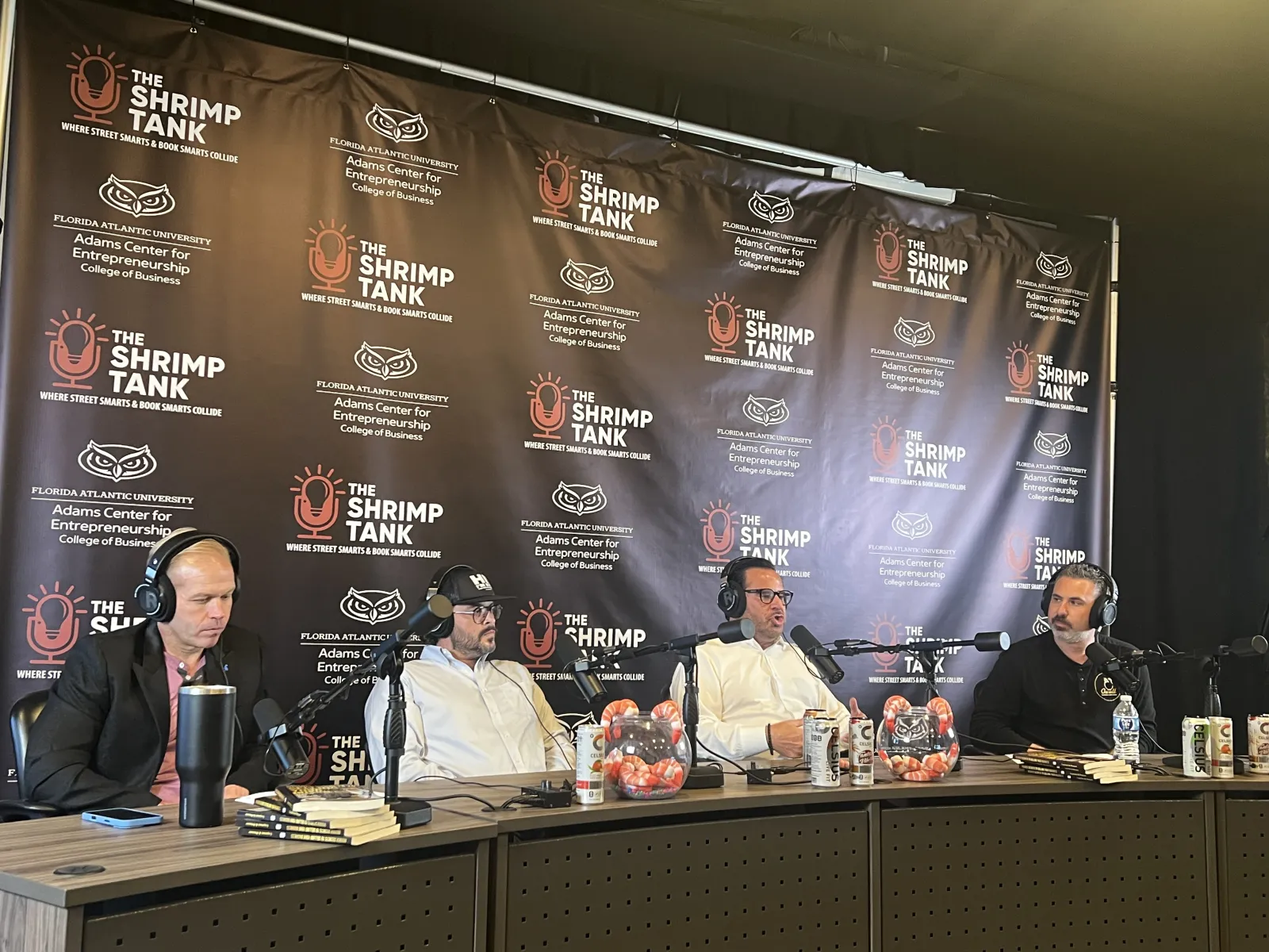 Four men wearing headsets engaged in a podcast discussion in front of The Shrimp Tank banner at Florida Atlantic University.