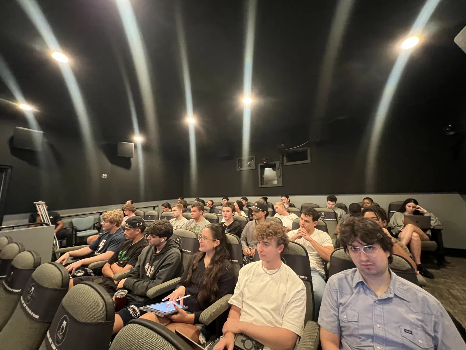 Group of students attentively seated in a dark lecture hall with black walls and ceiling lights.