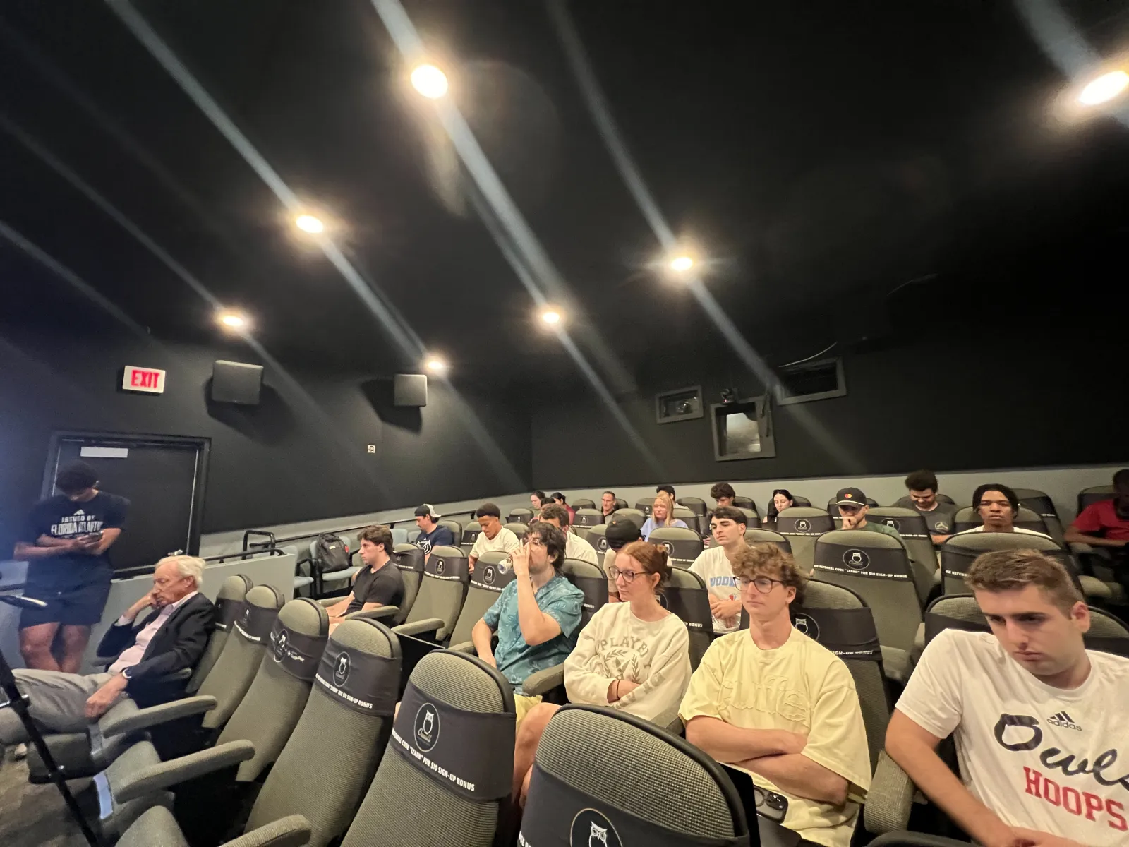 Group of people seated in a small, dimly lit theater or lecture room with black walls and ceiling lights.