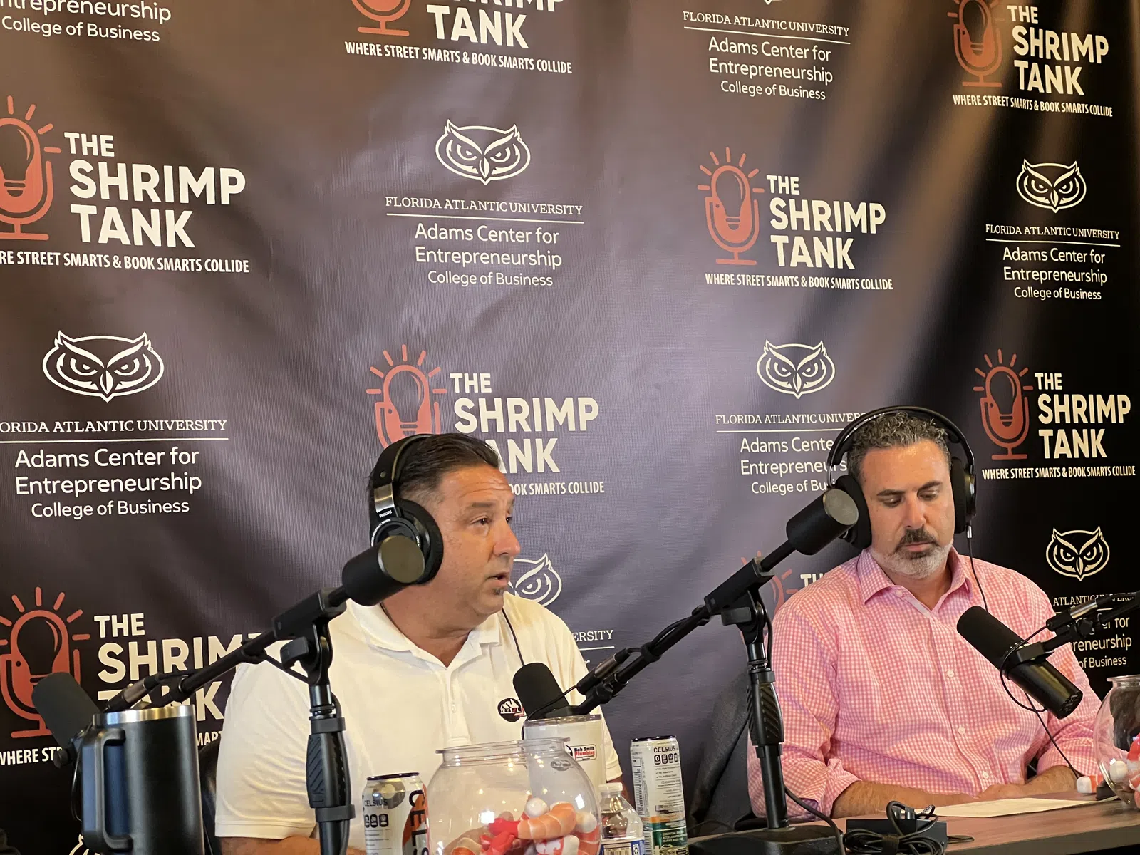 Two men wearing headphones speaking into microphones during The Shrimp Tank podcast recording with branded backdrop
