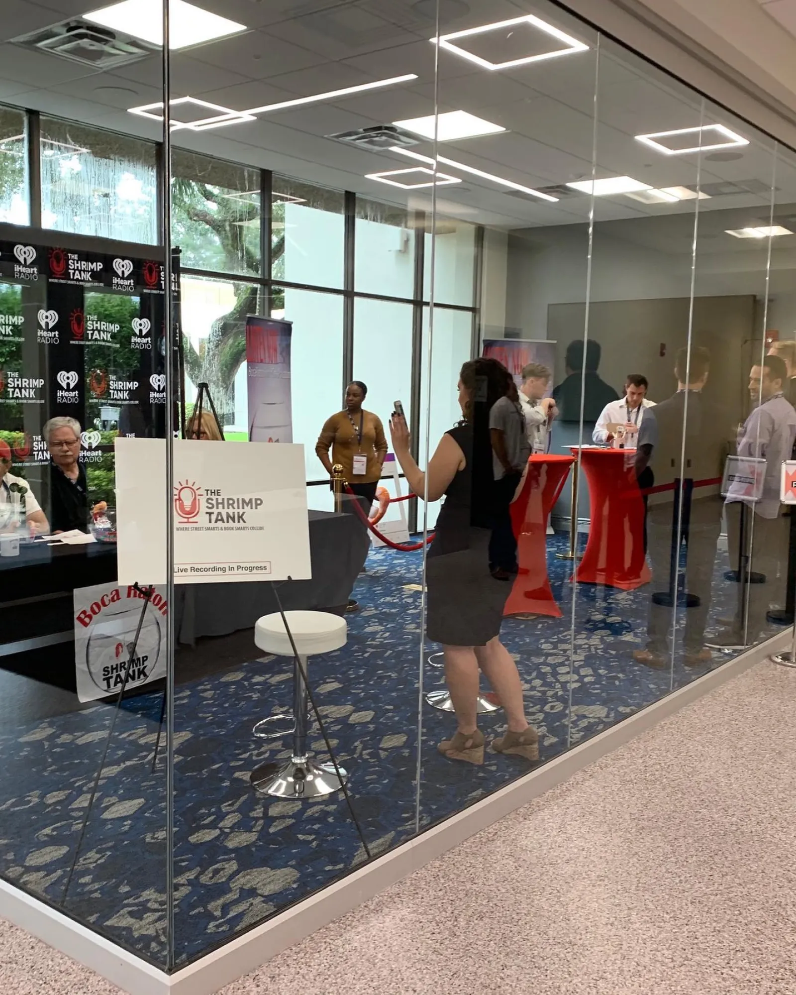People attending a live recording of The Shrimp Tank podcast in a glass-walled conference room with banners and tables.