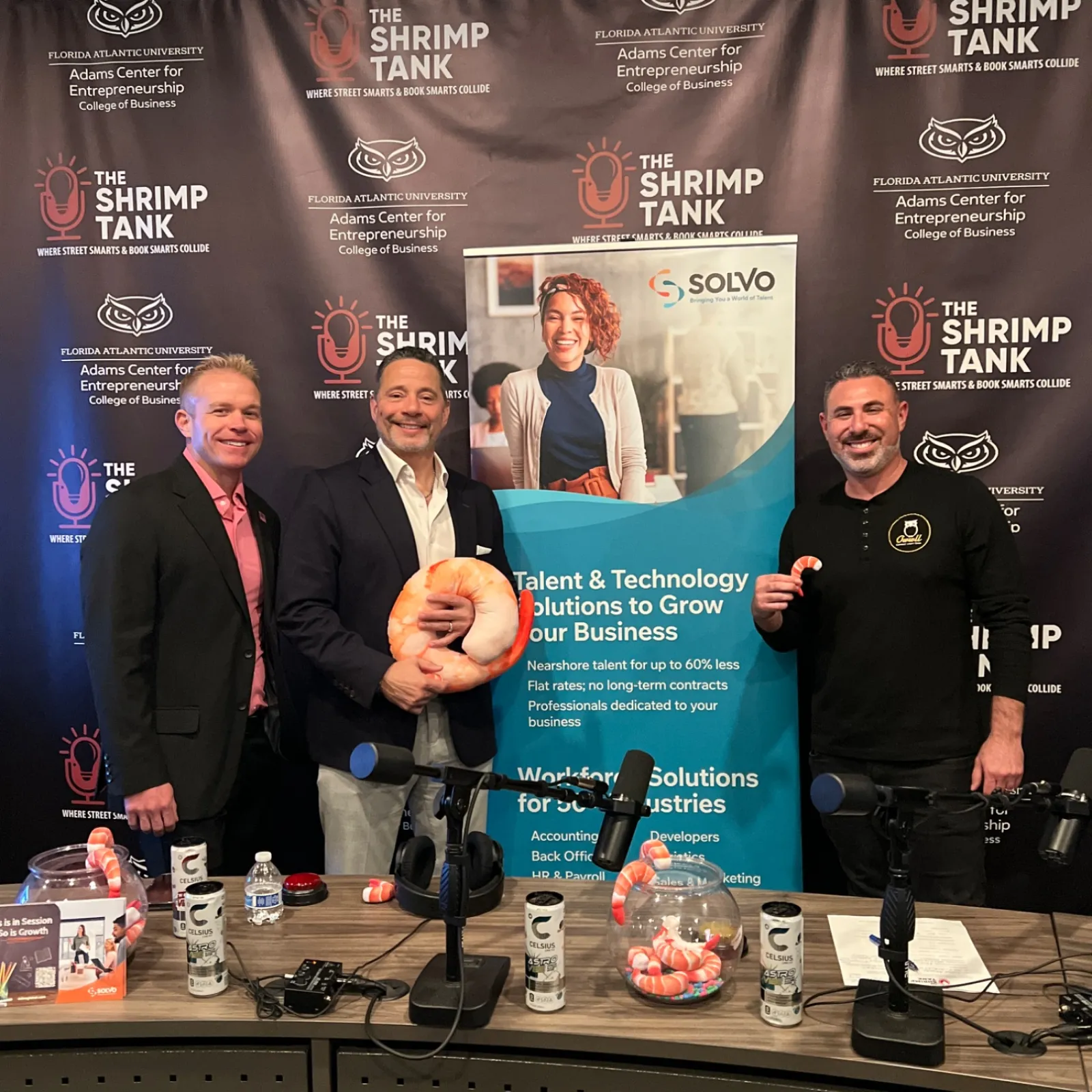 Three men posing with shrimp props in front of The Shrimp Tank podcast backdrop with promotional materials on table.