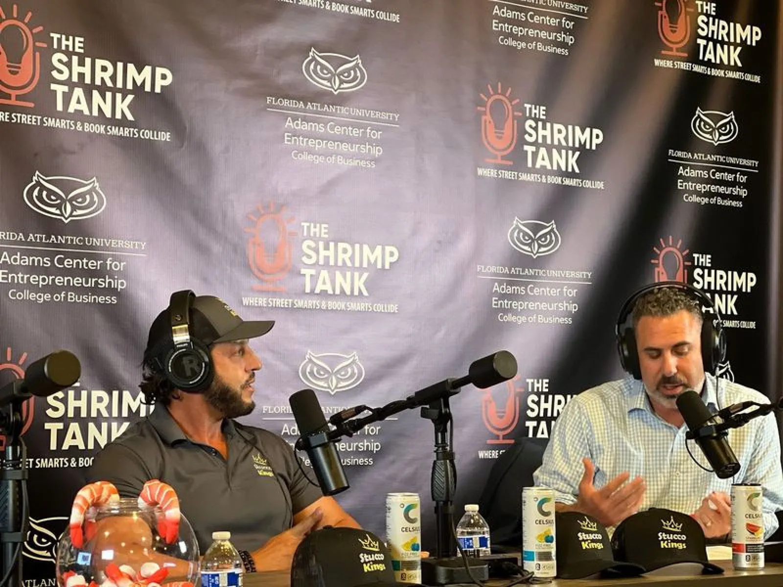 Two men wearing headphones speaking into microphones at The Shrimp Tank podcast backdrop with drinks and hats on the table