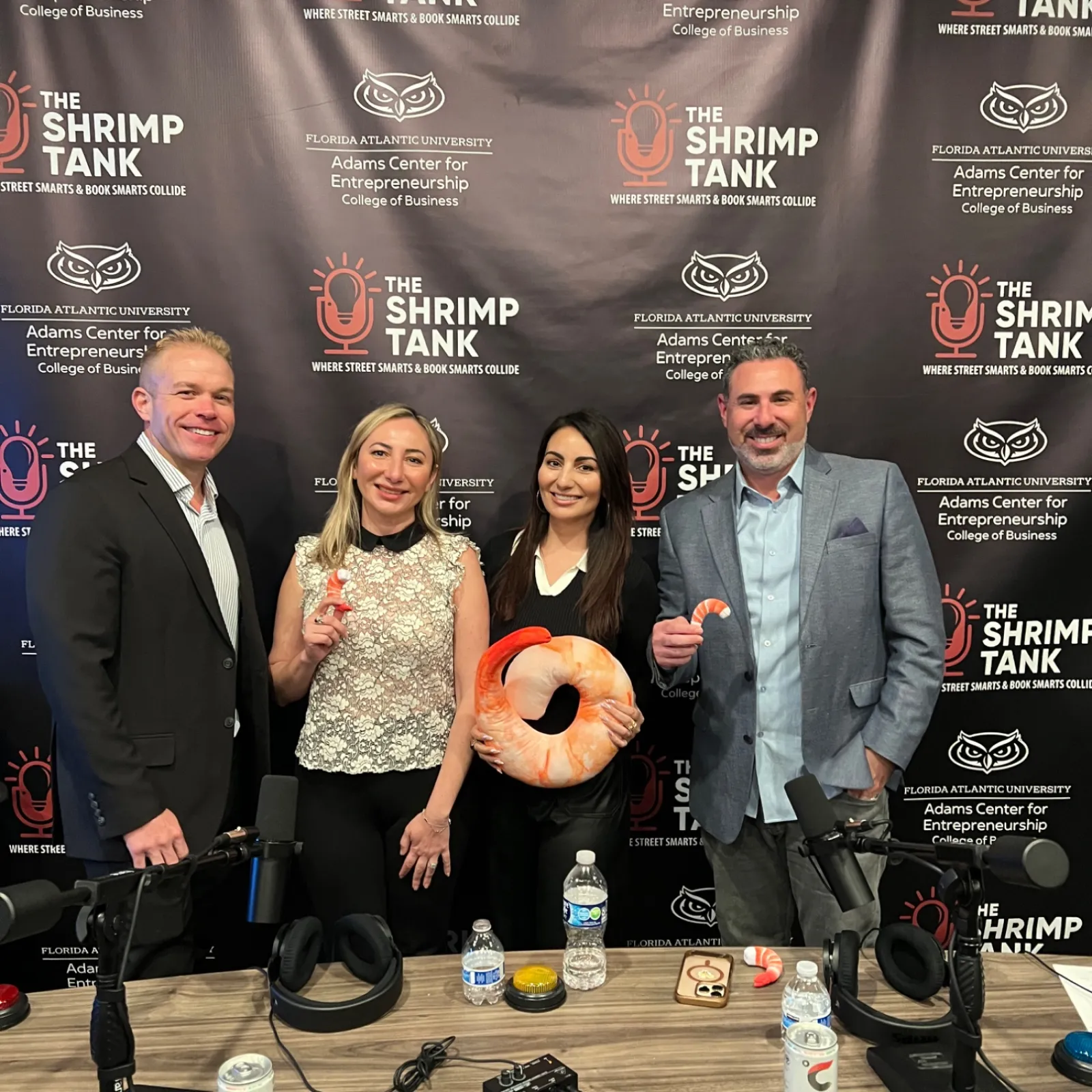 Four business professionals pose in front of The Shrimp Tank branded backdrop holding shrimp-themed items at a podcast setup.