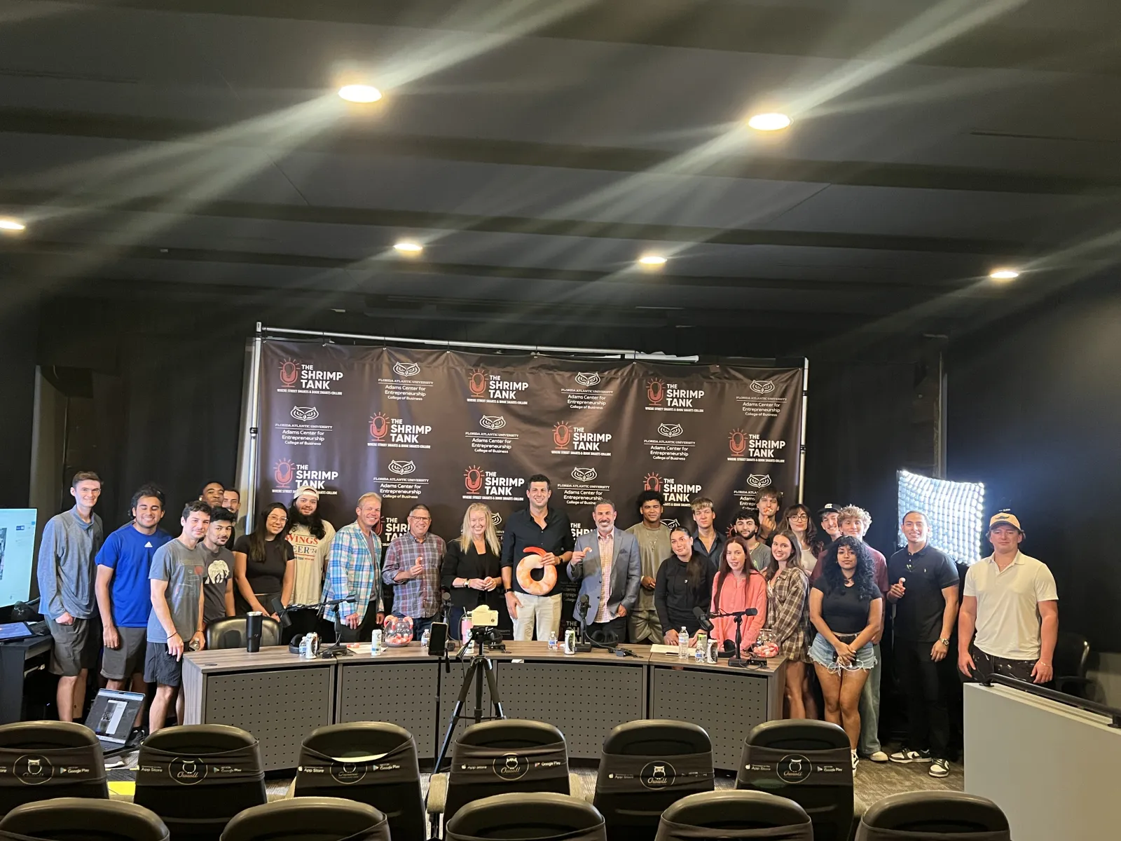 Group photo of diverse people standing behind a table with a Shrimp Tank backdrop and bright studio lights