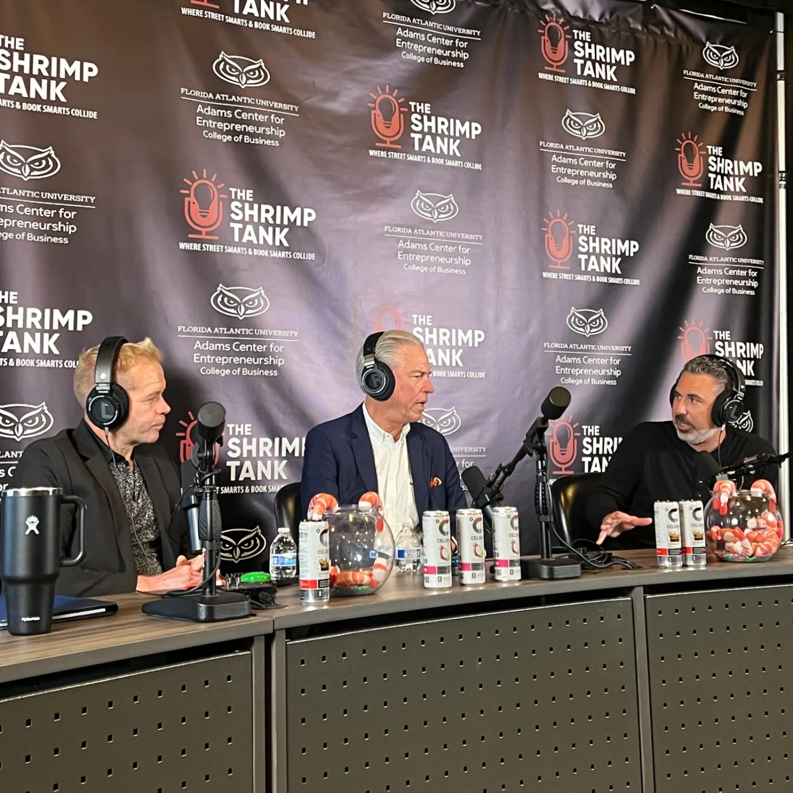 Three men wearing headphones sit at a podcast table with microphones and a The Shrimp Tank branded backdrop.