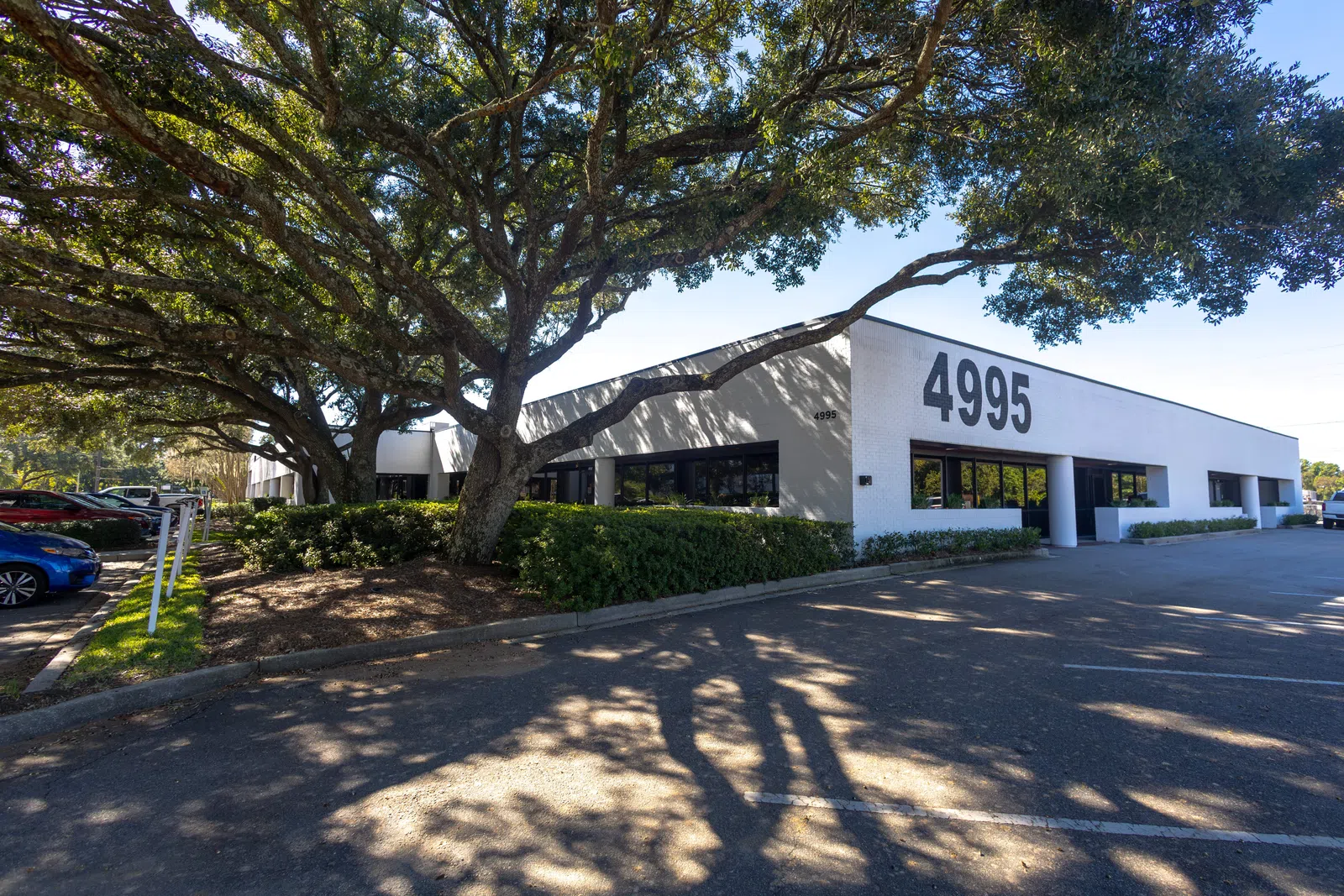 Modern low-rise office building with large '4995' numbers, shaded by large oak trees on a sunny day