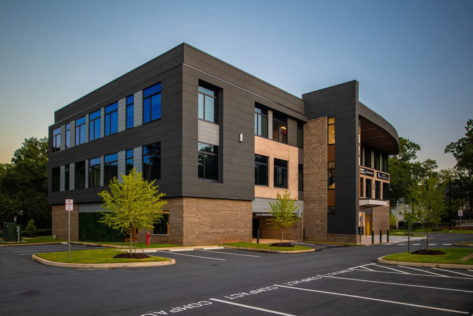 Modern two-story office building with black and brick exterior surrounded by trees and empty parking lot at dusk