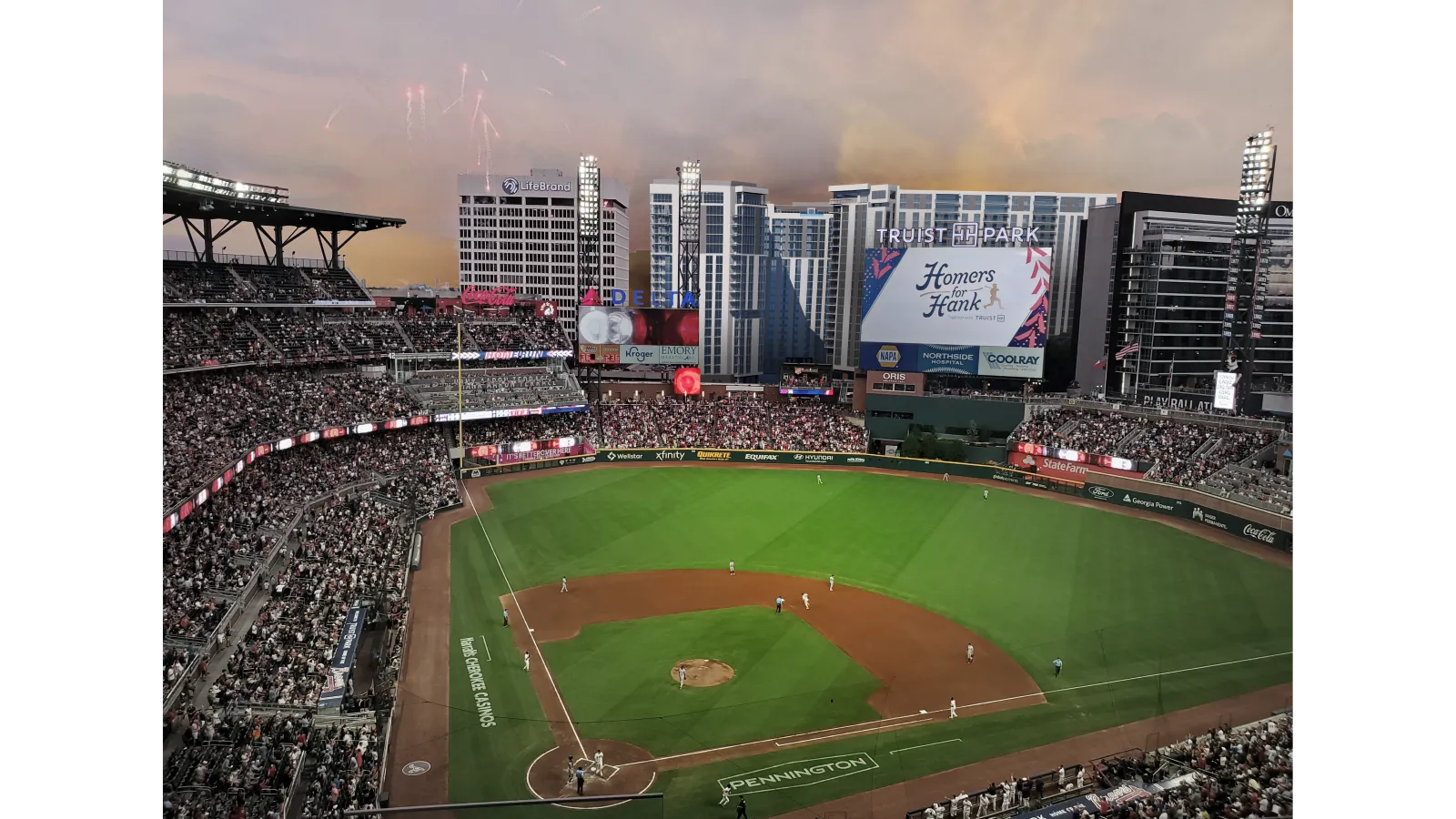 Twilight view of a packed baseball stadium with players on field and fireworks in sky.