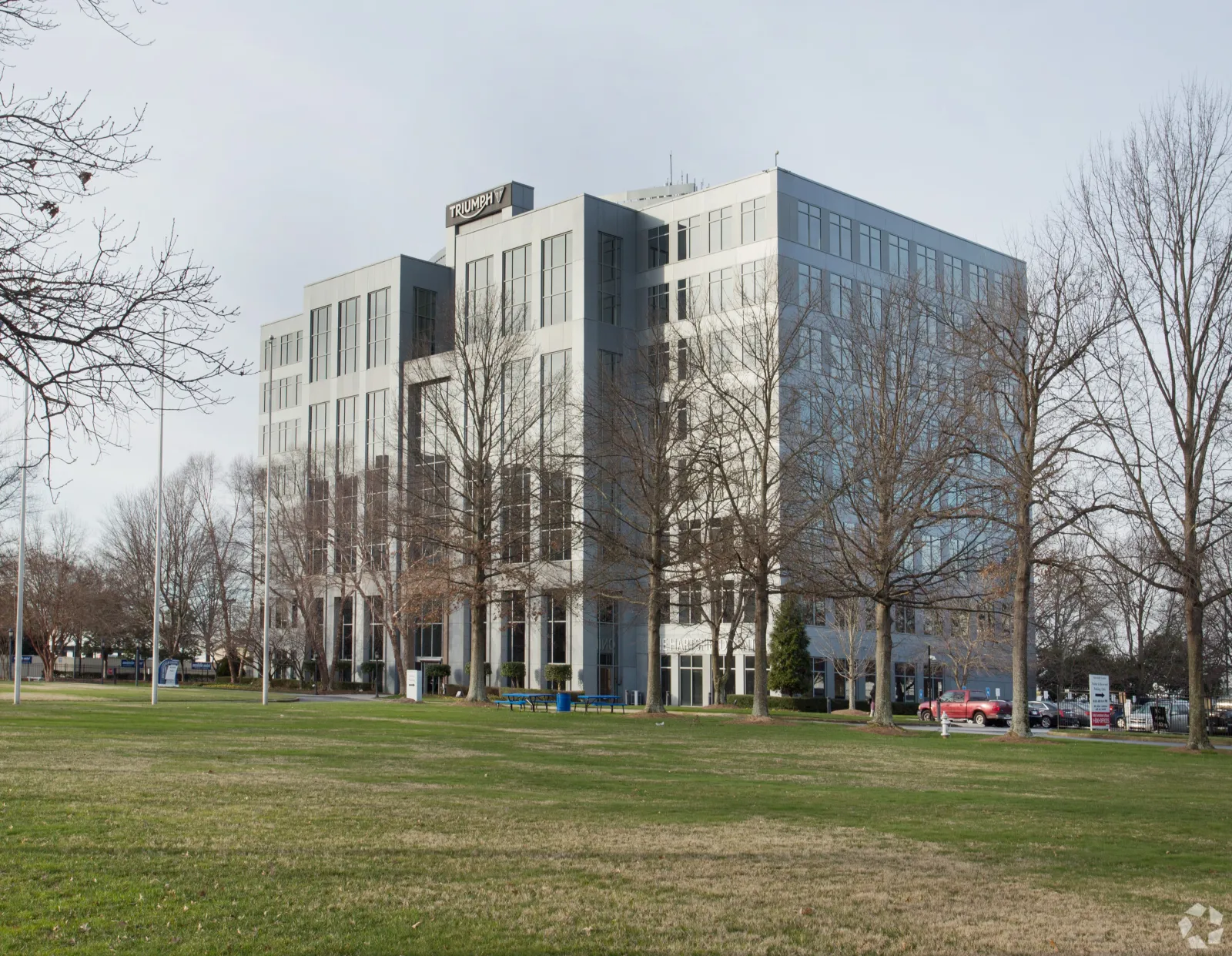 Modern office building with glass windows behind leafless trees and a green lawn under cloudy sky