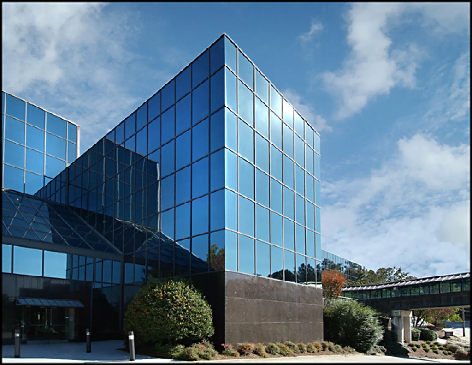 Modern glass office building reflecting blue sky with white clouds and surrounding greenery.