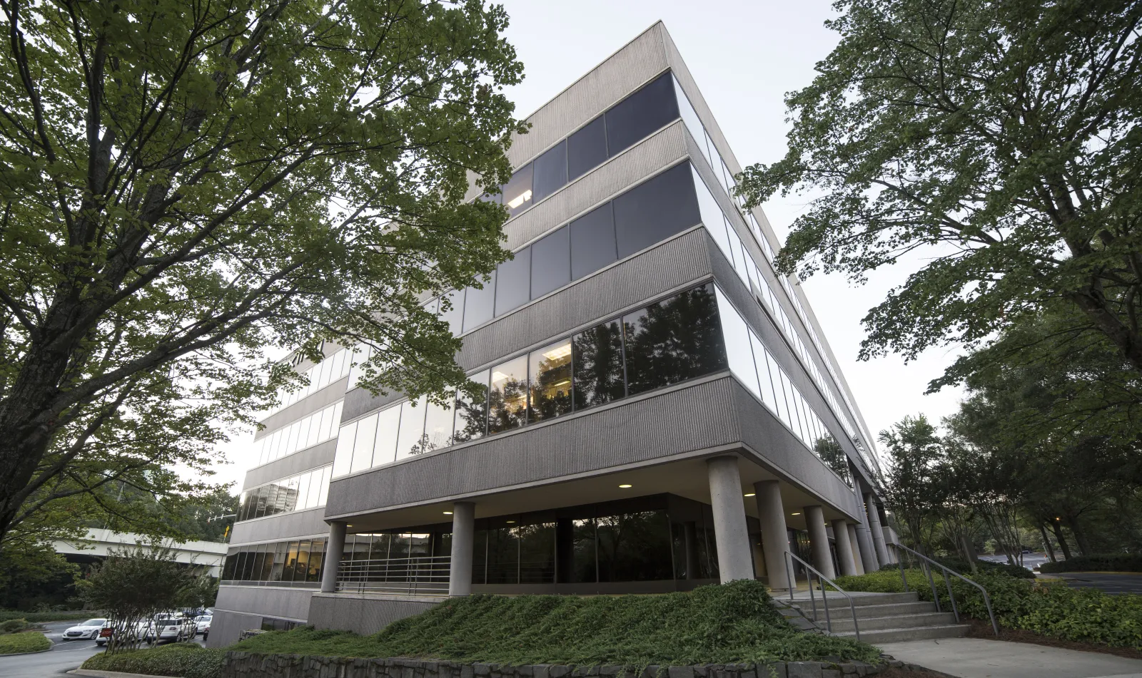 Modern office building surrounded by green trees with reflective windows and a sidewalk entrance.