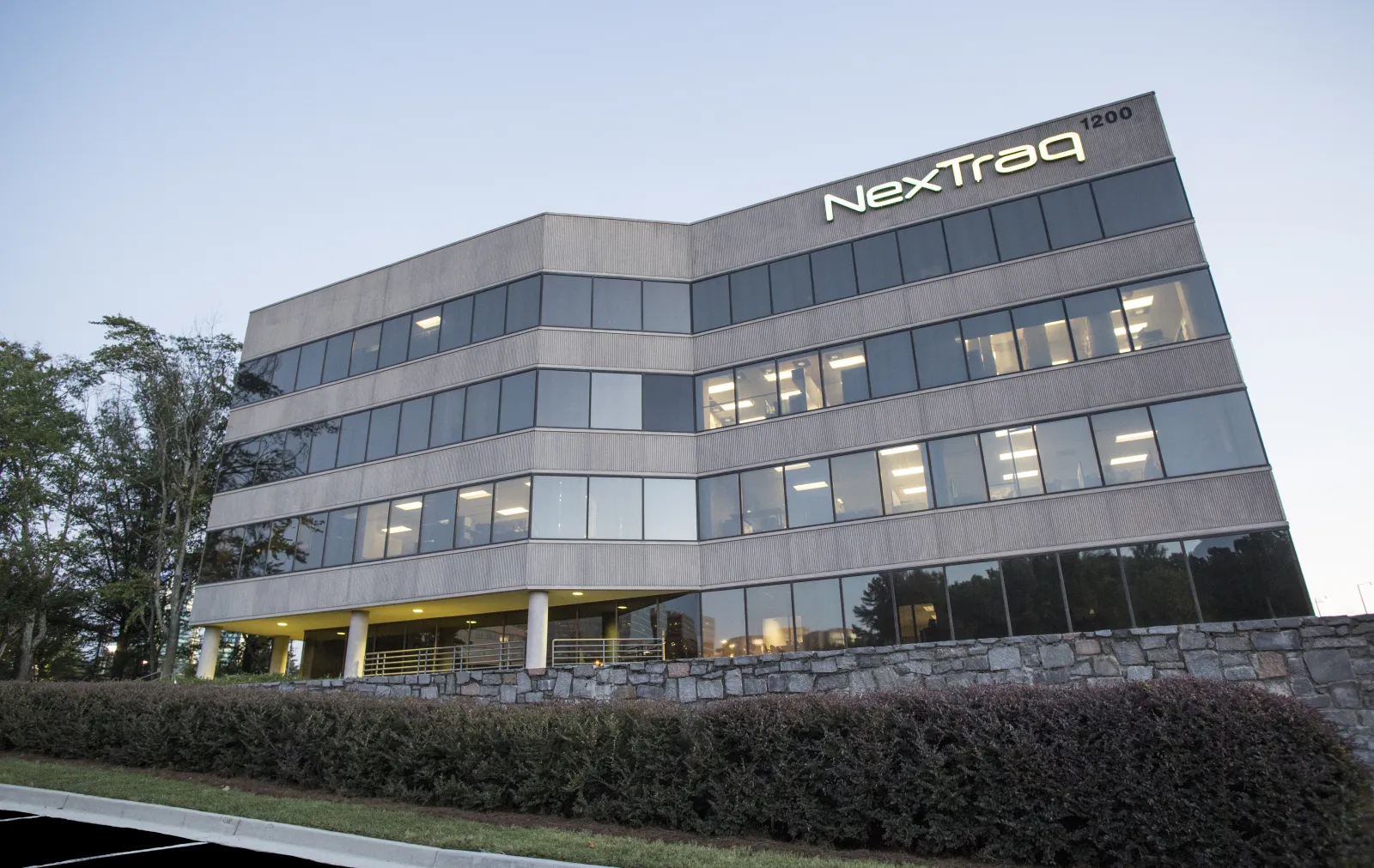 Modern office building with large windows and NextTraq logo at dusk surrounded by greenery and stone wall.