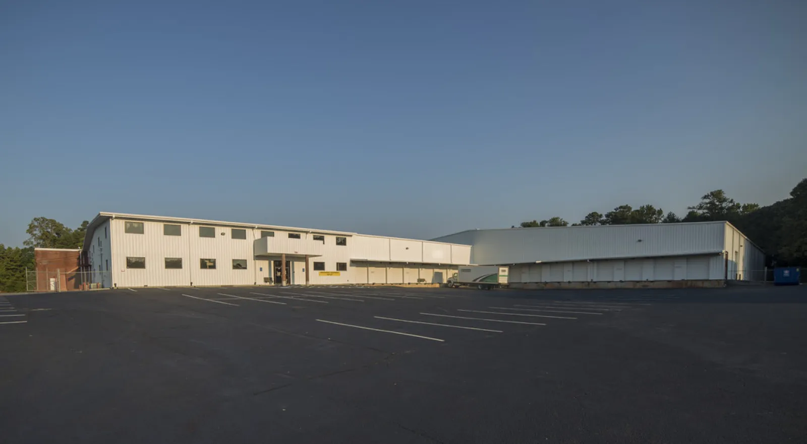 Large white industrial warehouse with empty parking lot under clear blue sky in daylight.