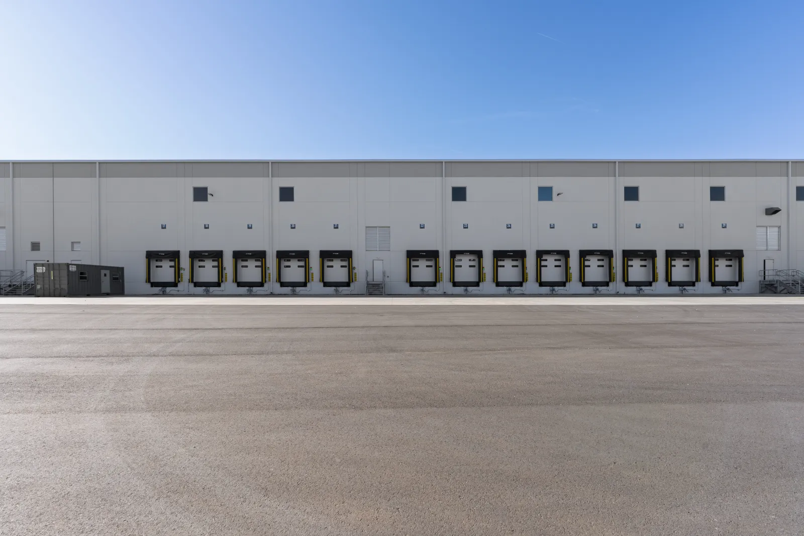Warehouse exterior with multiple loading dock doors under a clear blue sky and empty asphalt foreground
