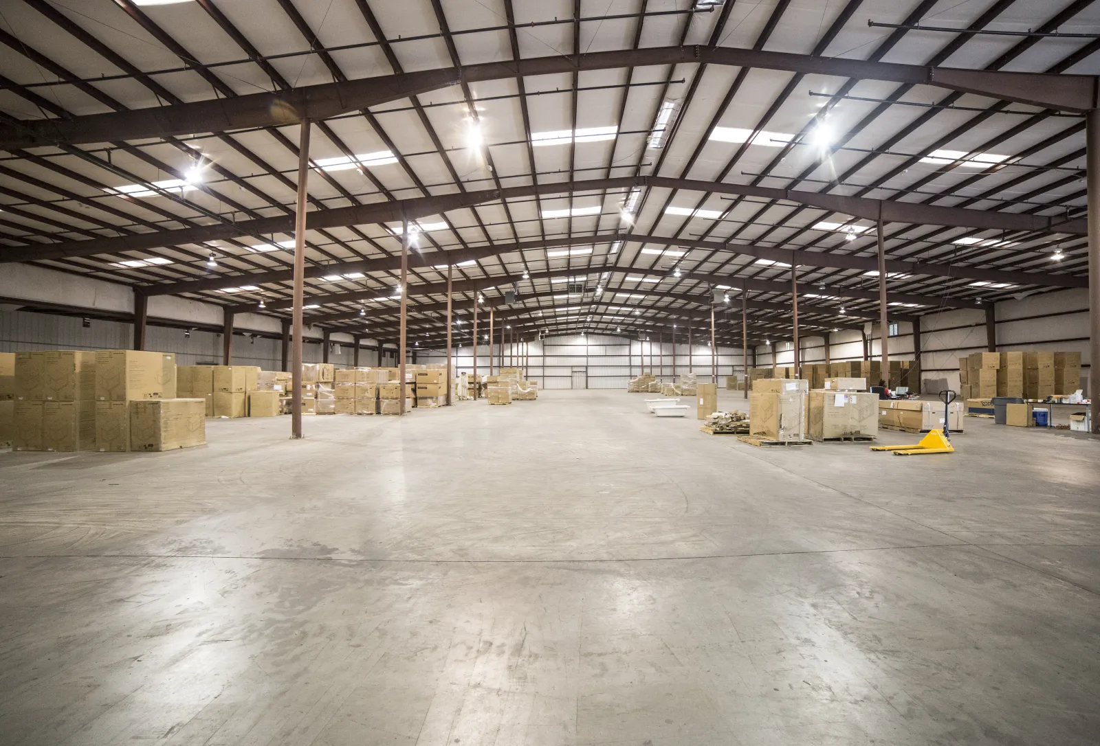 Spacious warehouse interior with concrete floor, metal beams, and stacks of cardboard boxes under bright lighting.