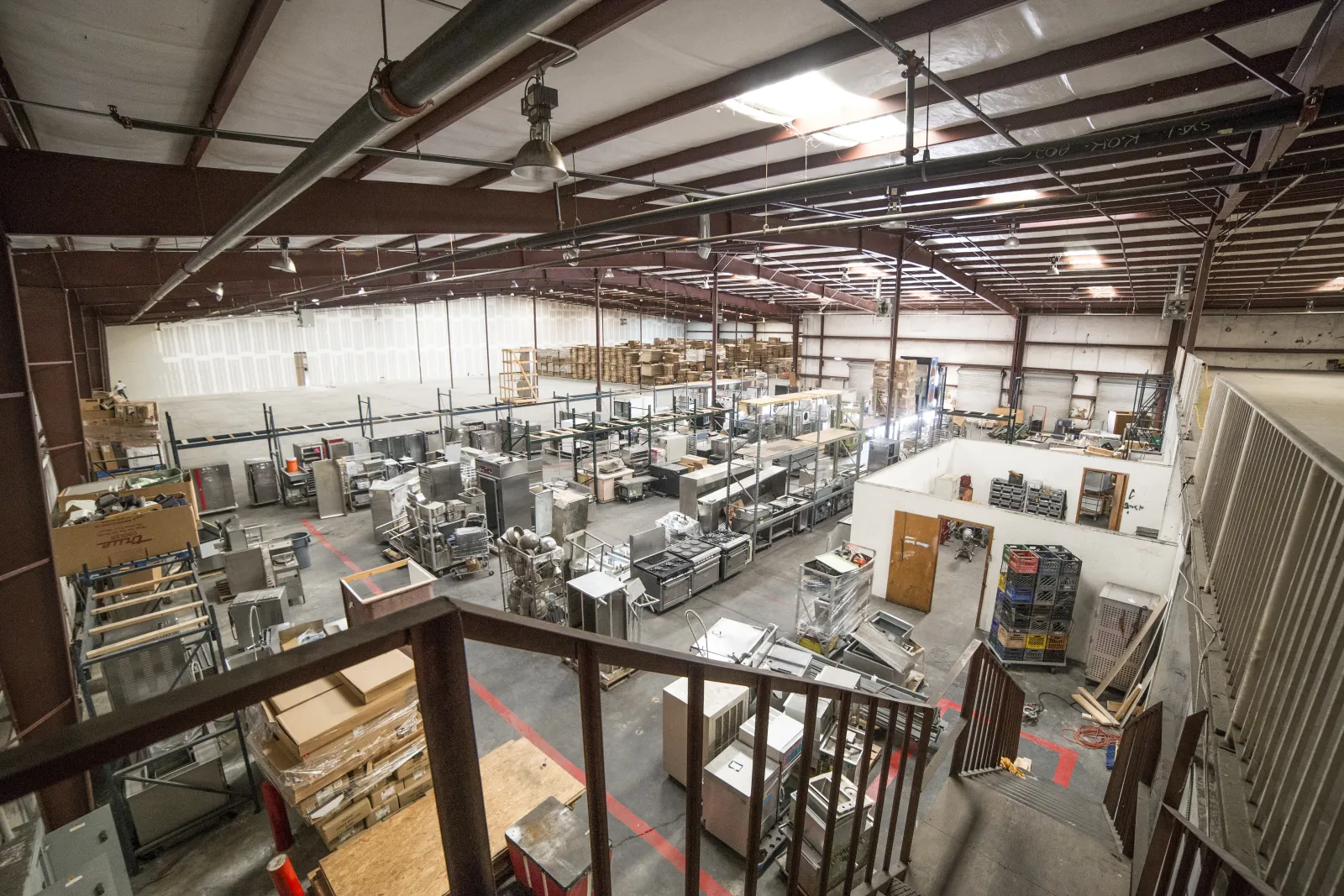 Spacious warehouse interior with industrial shelving, equipment, and boxes organized under a metal roof.