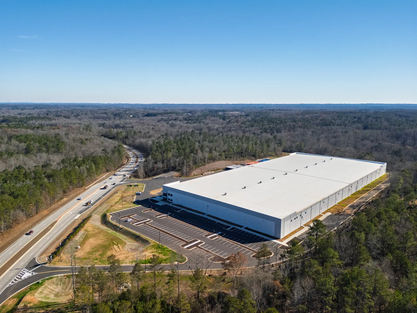 Aerial view of large white industrial warehouse surrounded by forest and adjacent to a road under clear sky