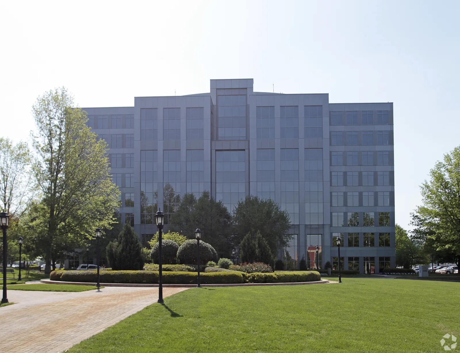 Modern glass office building with landscaped garden, trees, and lamp posts in the foreground on a sunny day