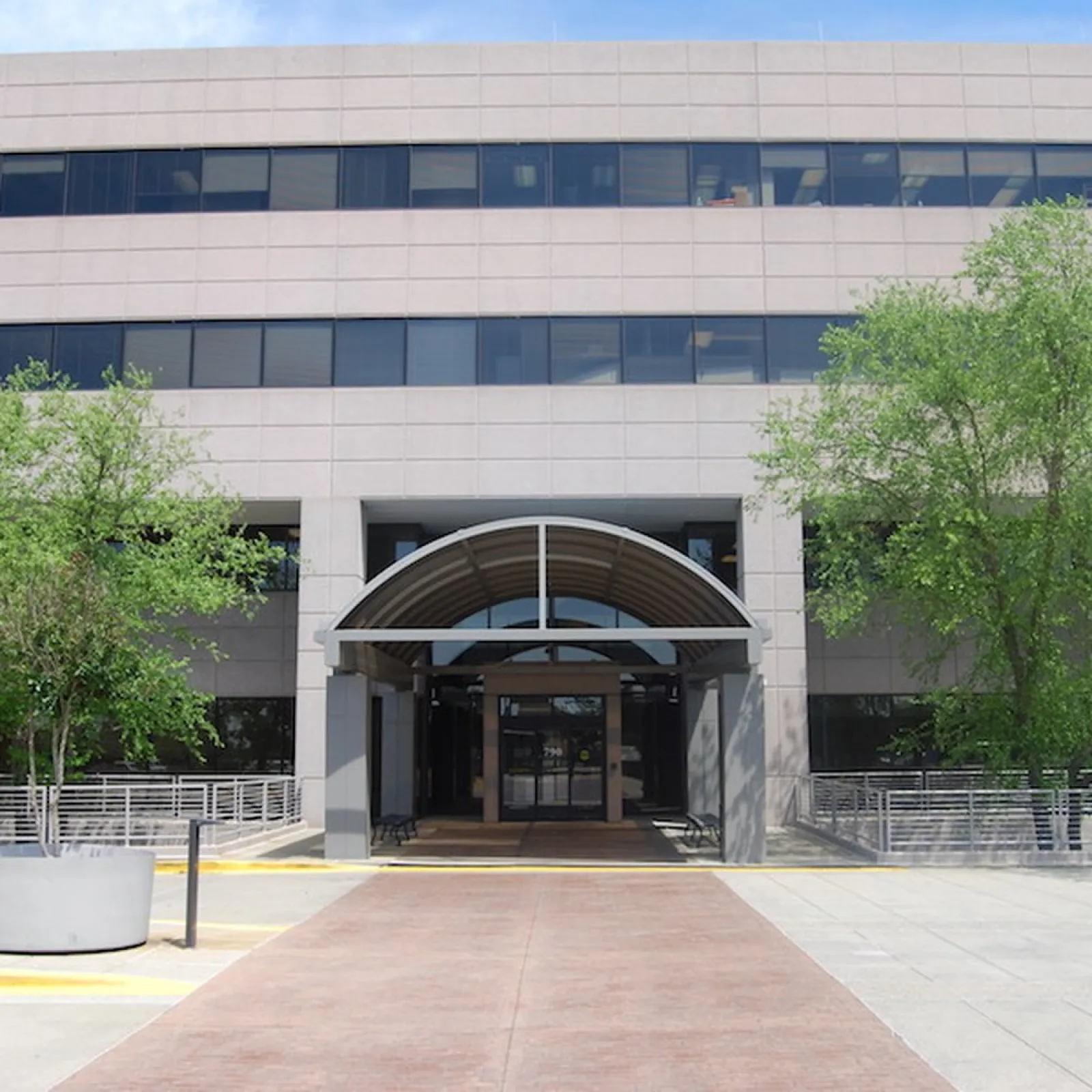 Modern office building entrance with arched canopy, surrounded by trees and concrete planters on a sunny day