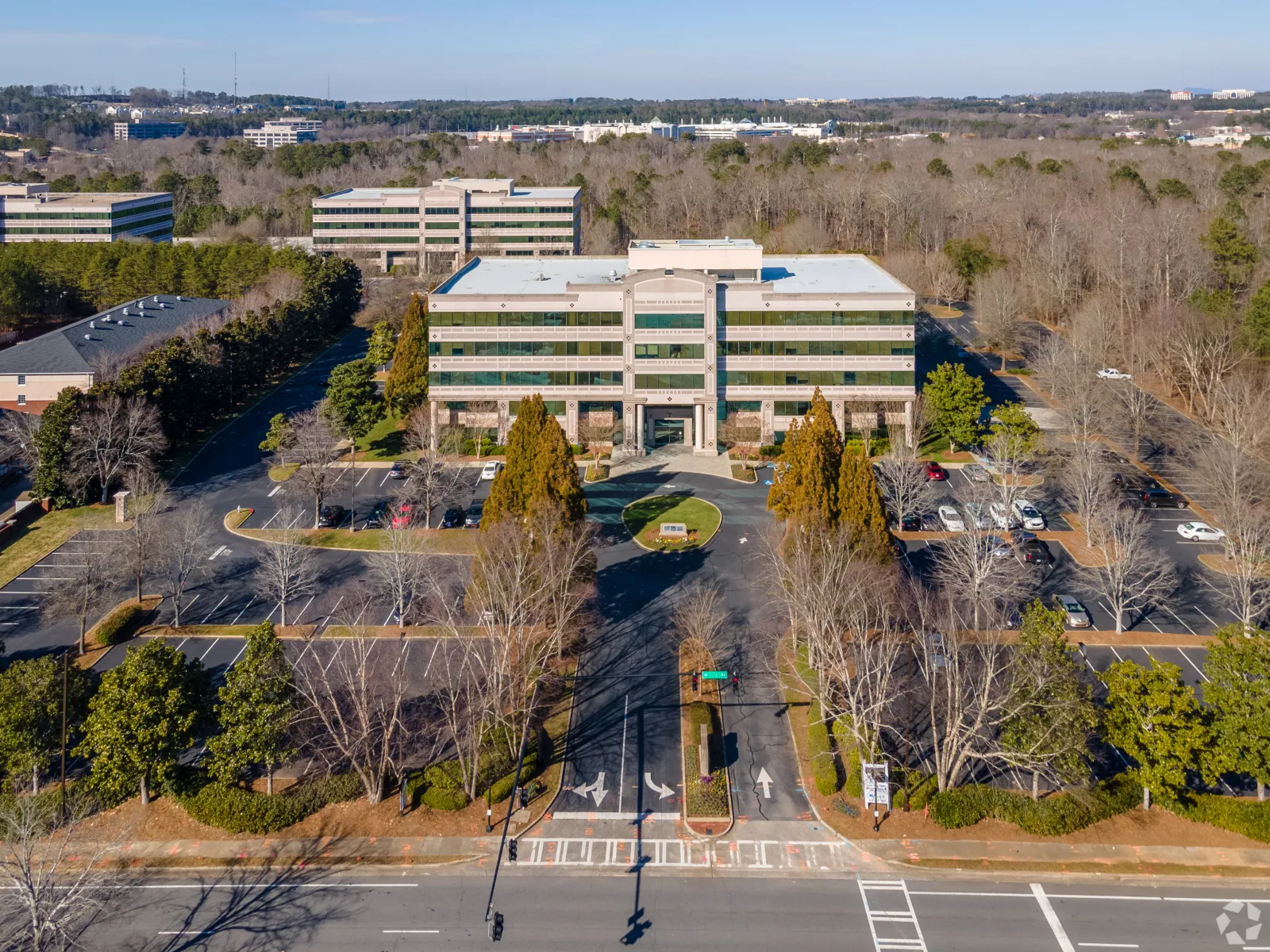 Aerial view of a modern office building surrounded by parking lots and trees in a suburban area under blue sky.