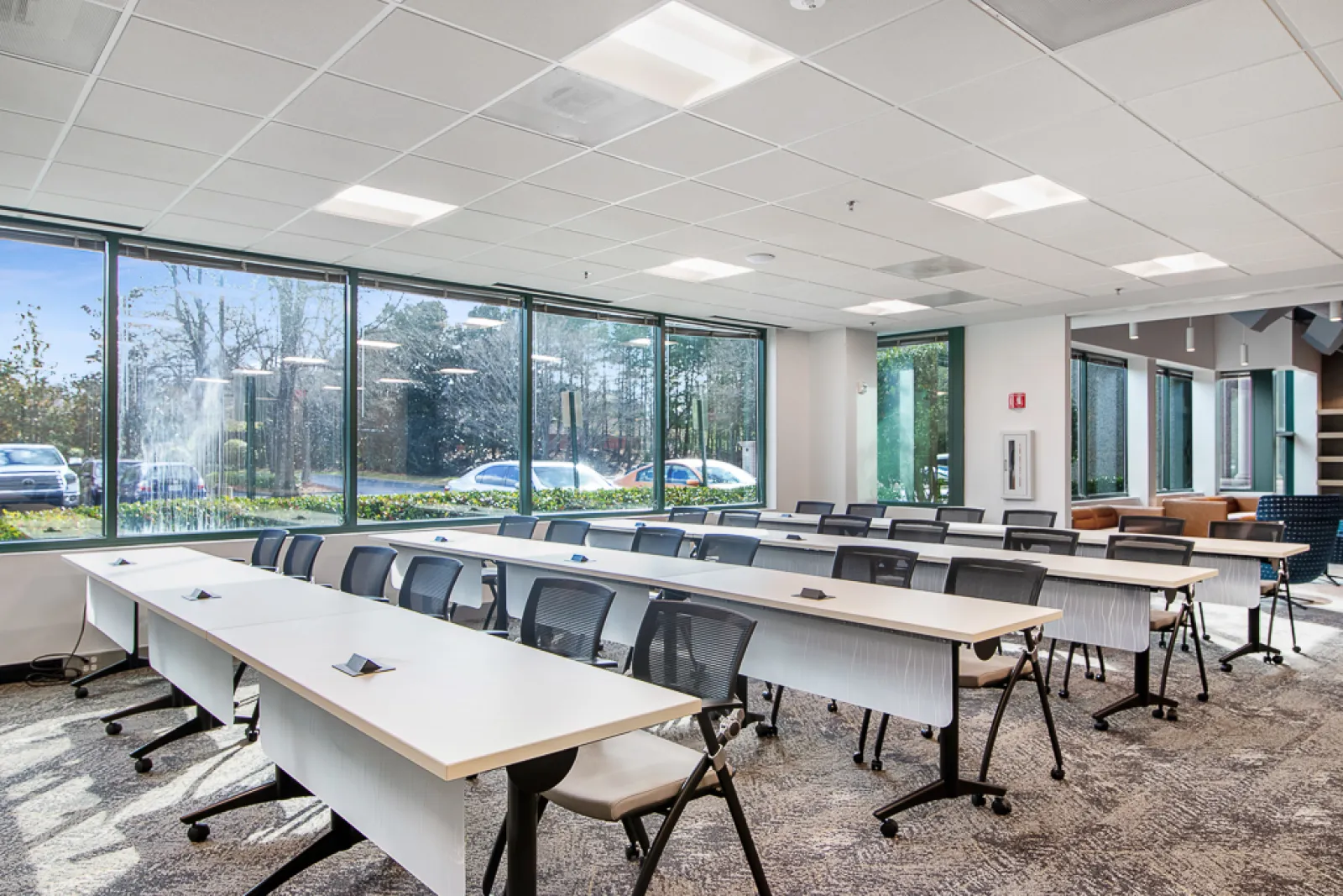 Bright modern conference room with large windows, rows of tables and black chairs, and carpeted floor.