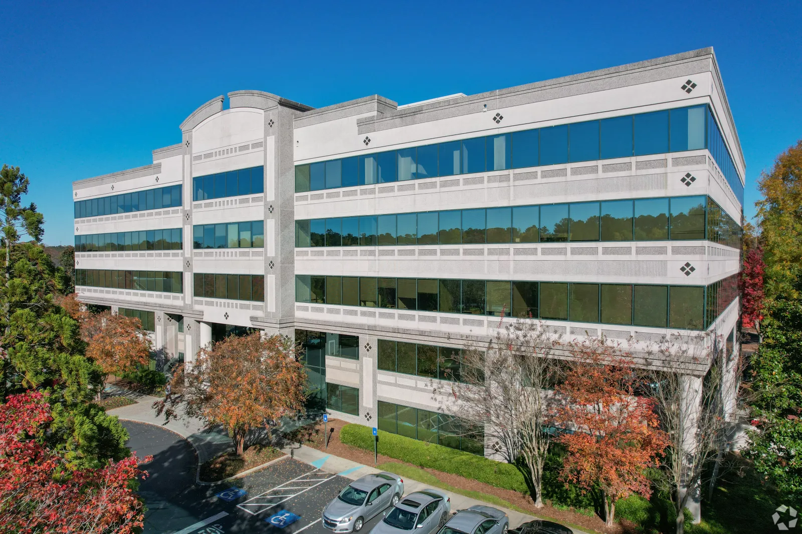 Modern four-story office building with large reflective windows surrounded by autumn trees and parked cars.