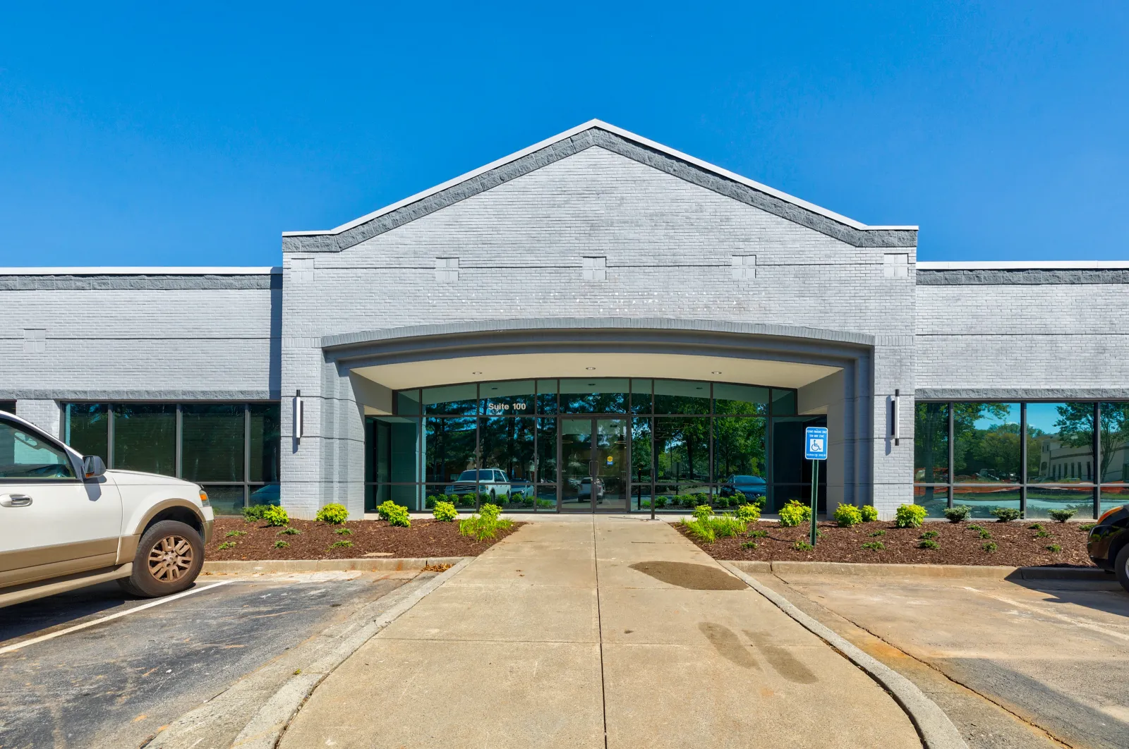 Modern gray brick commercial building entrance with glass doors and parking spaces under clear blue sky