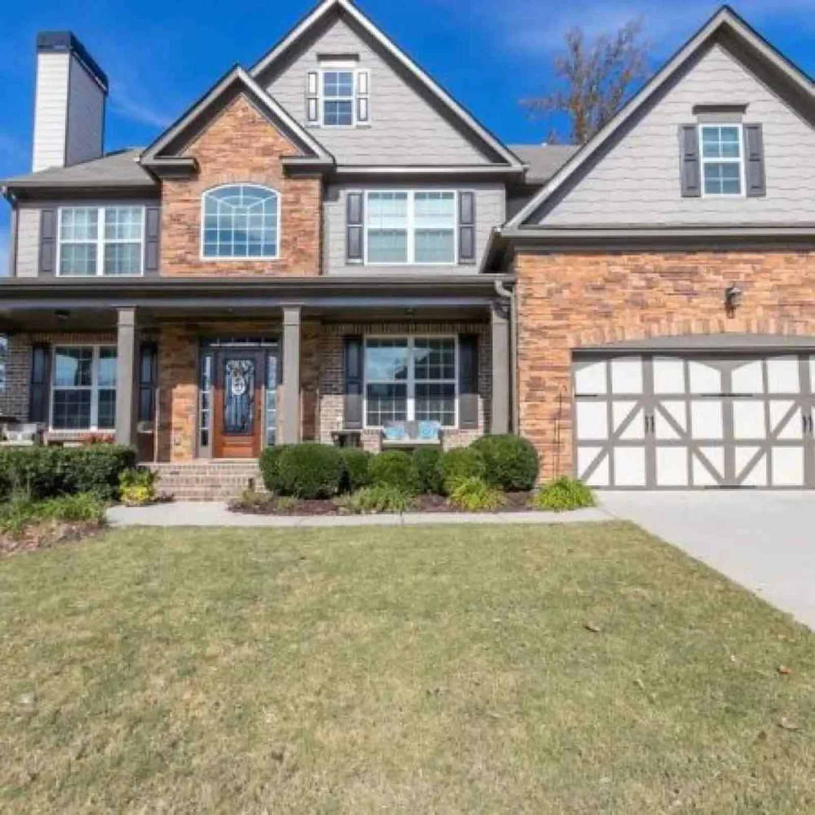 Two-story suburban house with brick and siding exterior, large windows, front porch, and a double garage under blue sky.