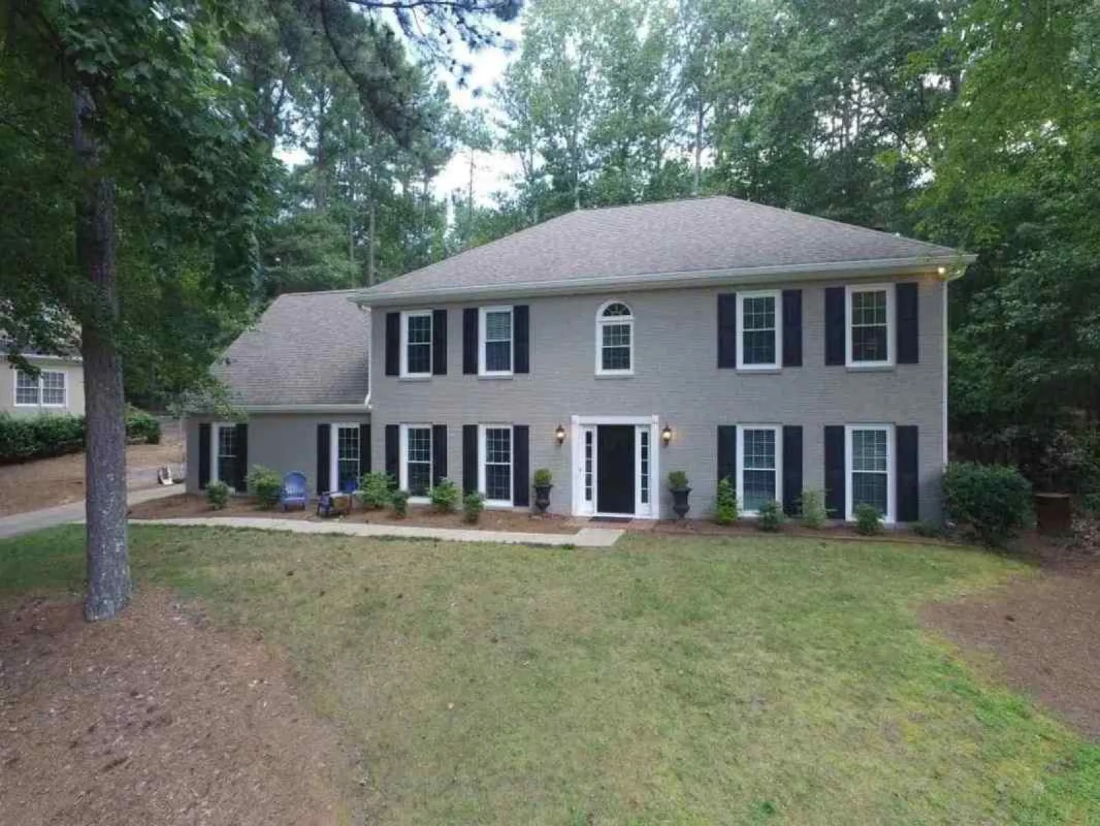 Two-story gray brick house with black shutters surrounded by trees and a spacious front yard.