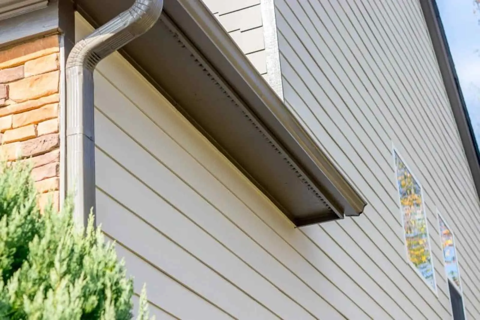 Close-up of house exterior showing beige vinyl siding, brown gutter system, and part of a brick wall with green shrub.