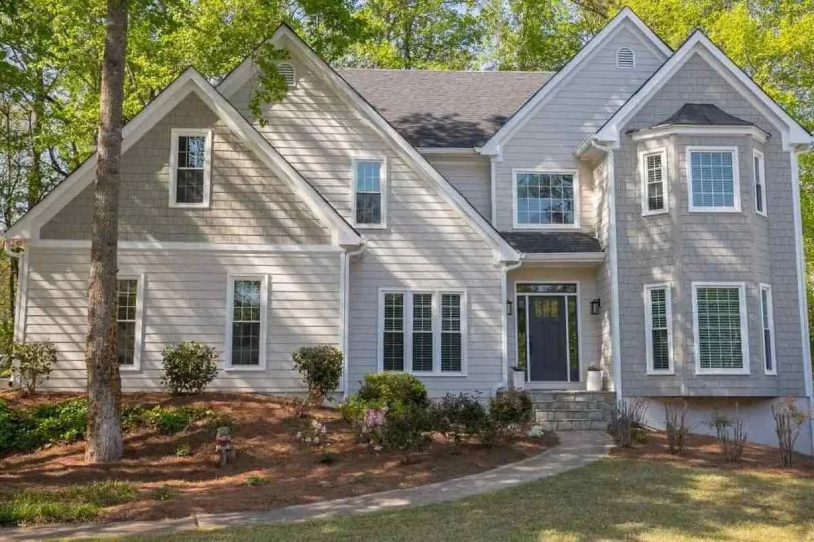 Two-story modern gray house with large windows surrounded by trees and landscaped garden under sunlight