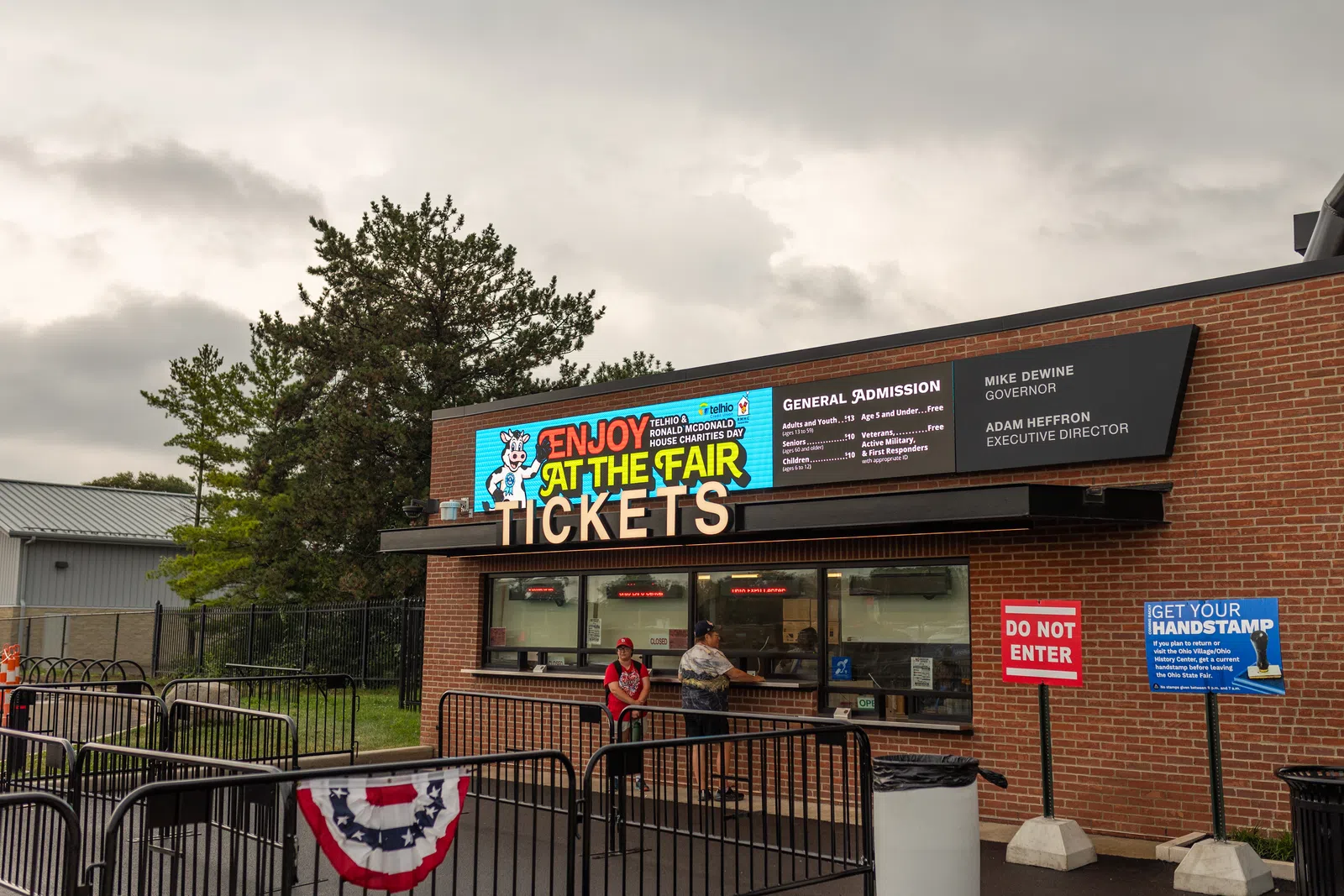 Ticket booth at the fair with 'Enjoy at the Fair' sign and general admission prices displayed on brick building.