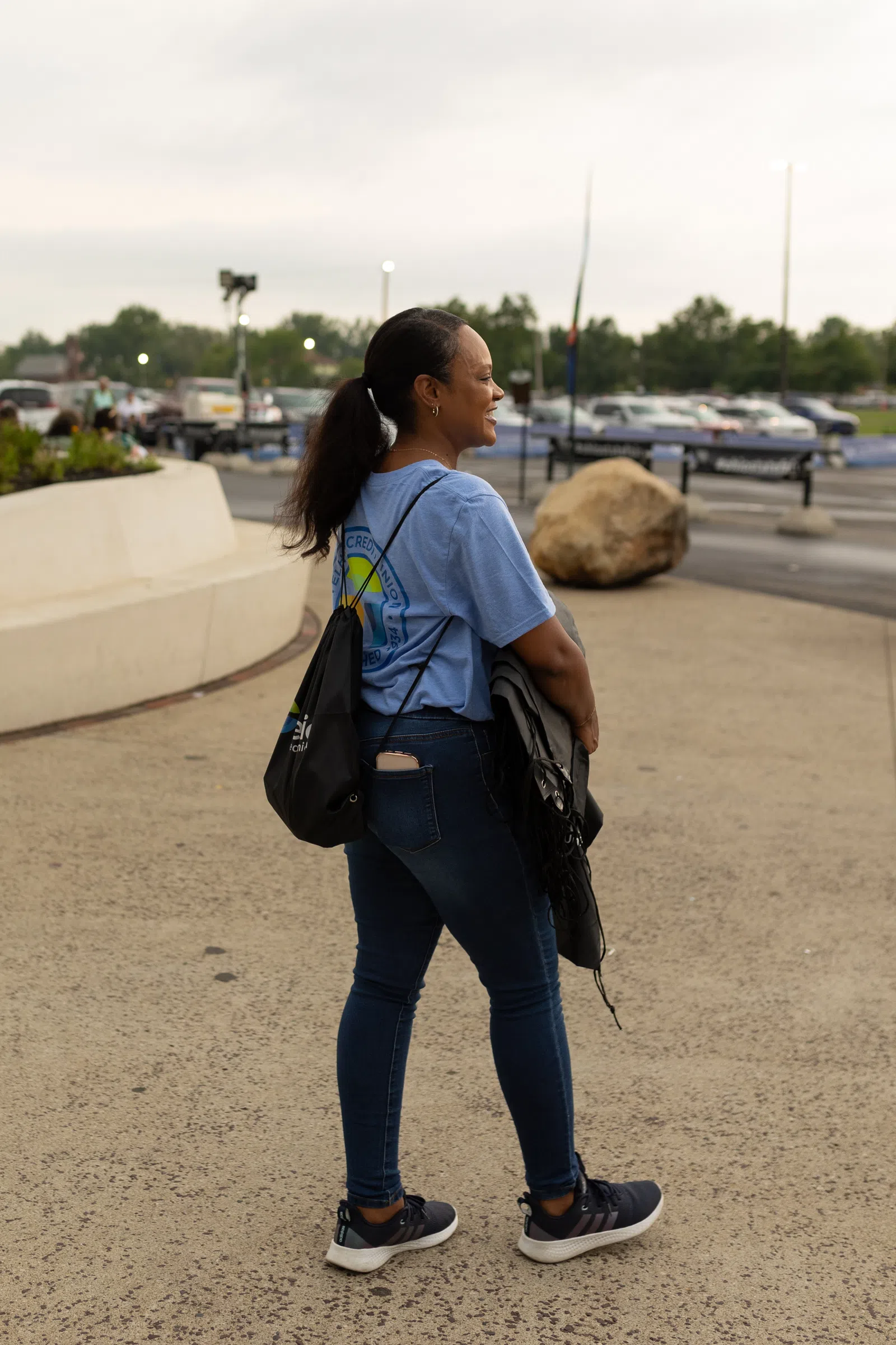 Woman in blue shirt and jeans standing outdoors with a black backpack and jacket, smiling and looking away.