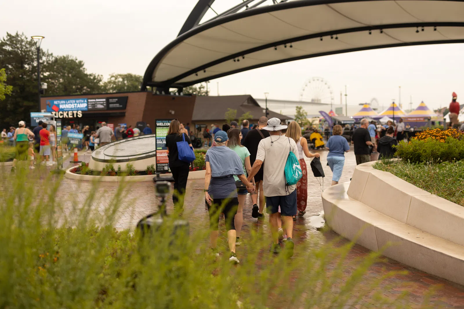 Crowd walking towards amusement park entrance with ferris wheel and colorful tents in the background.