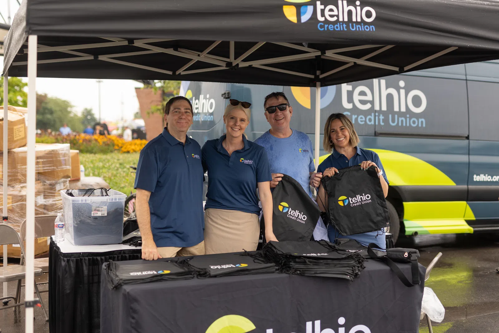 Group of four smiling people in Telhio Credit Union shirts at promotional booth with branded bags and van.