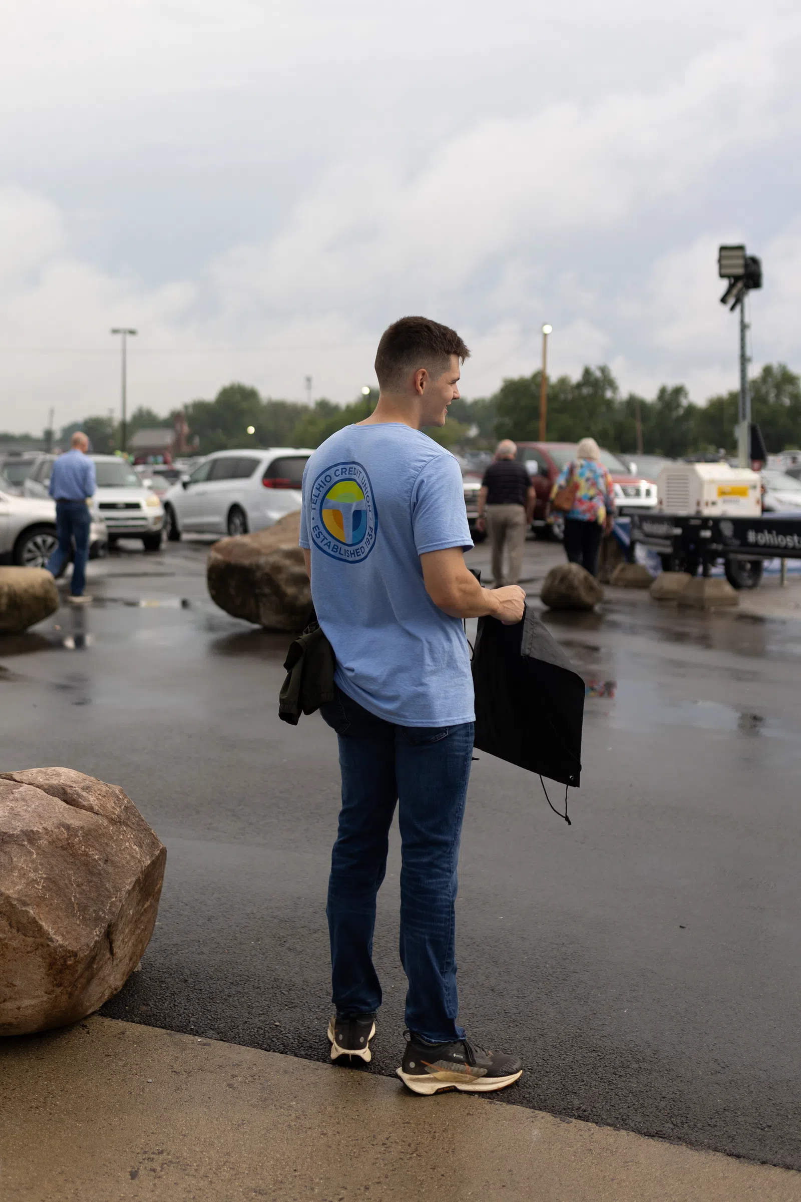 Young man in blue t-shirt and jeans holding a jacket standing in wet parking lot near large rocks and cars.