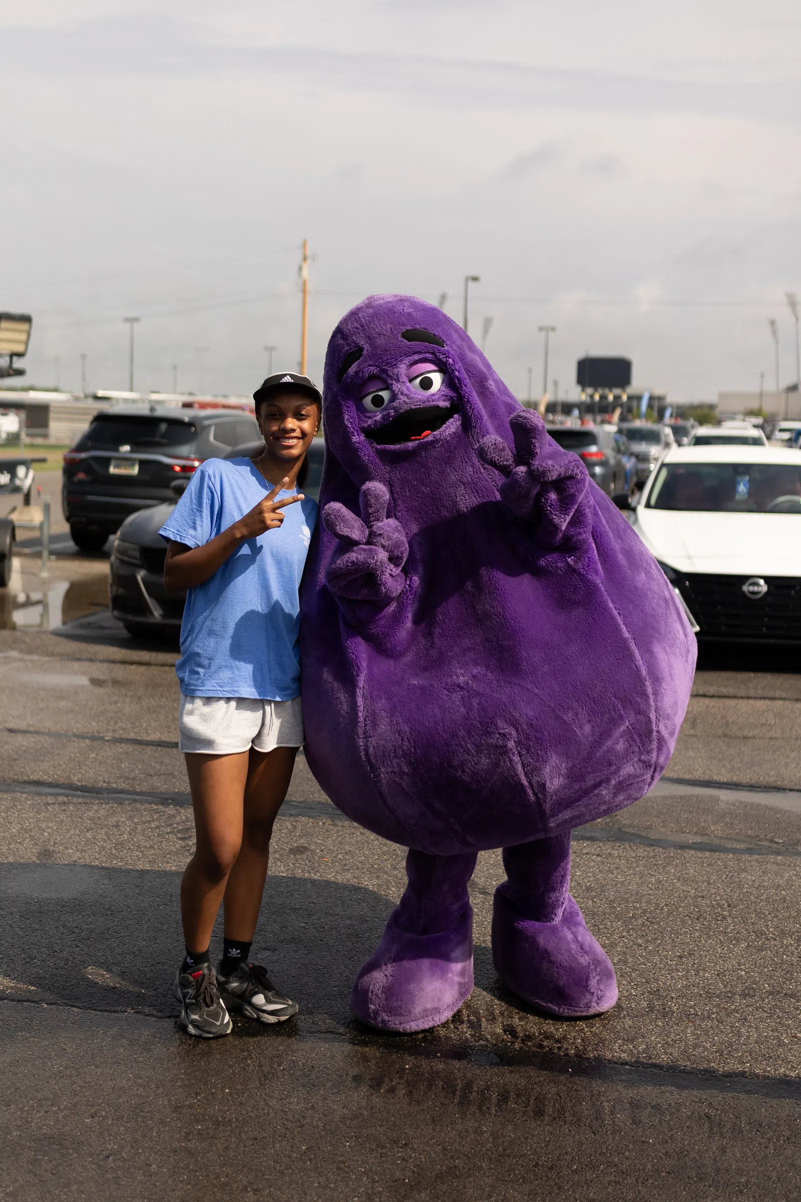 Young woman standing next to a large purple mascot posing with peace signs in a parking lot.