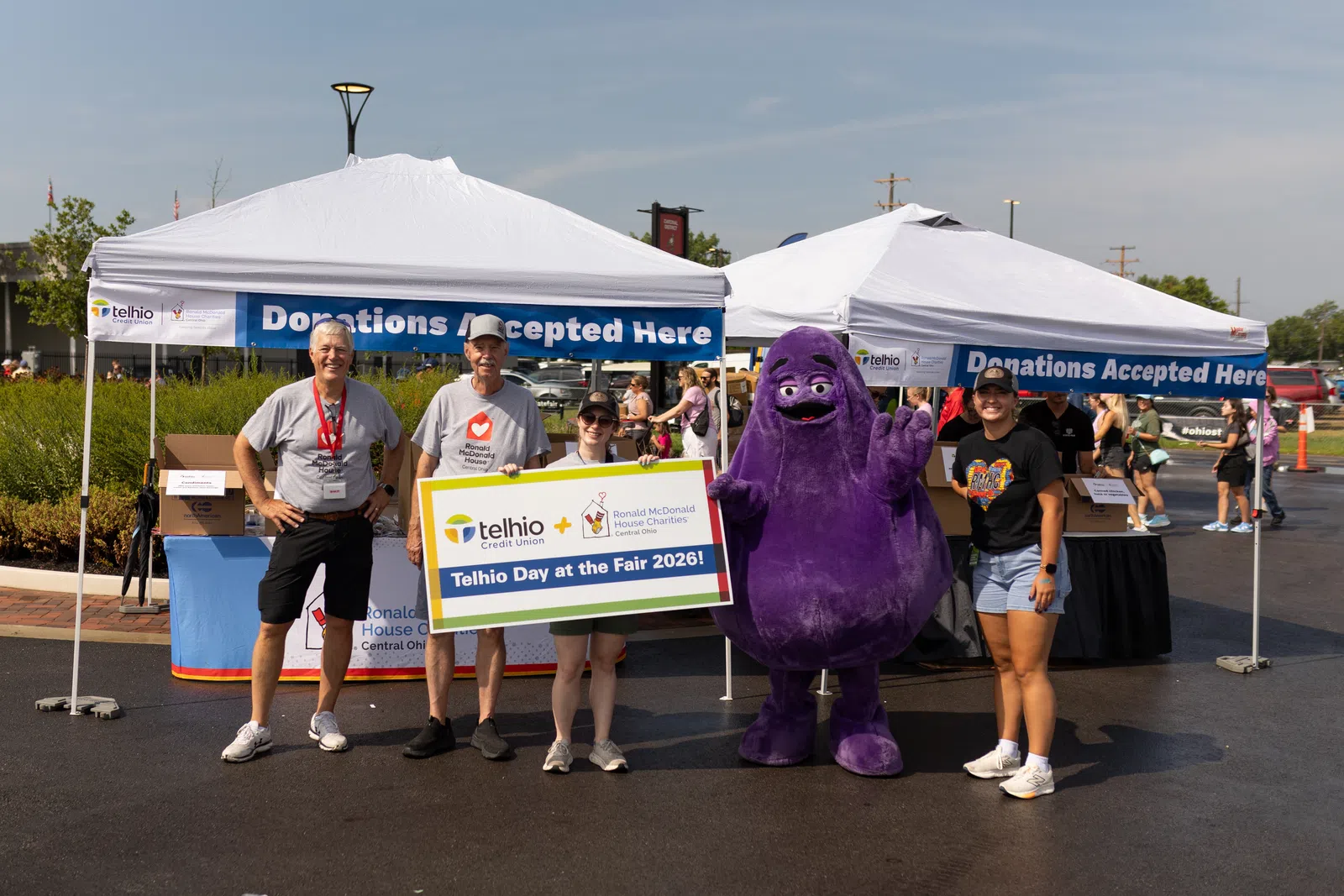 Group of people at donation tents holding a sign promoting Telhio Day at the Fair 2026 with a purple mascot character.