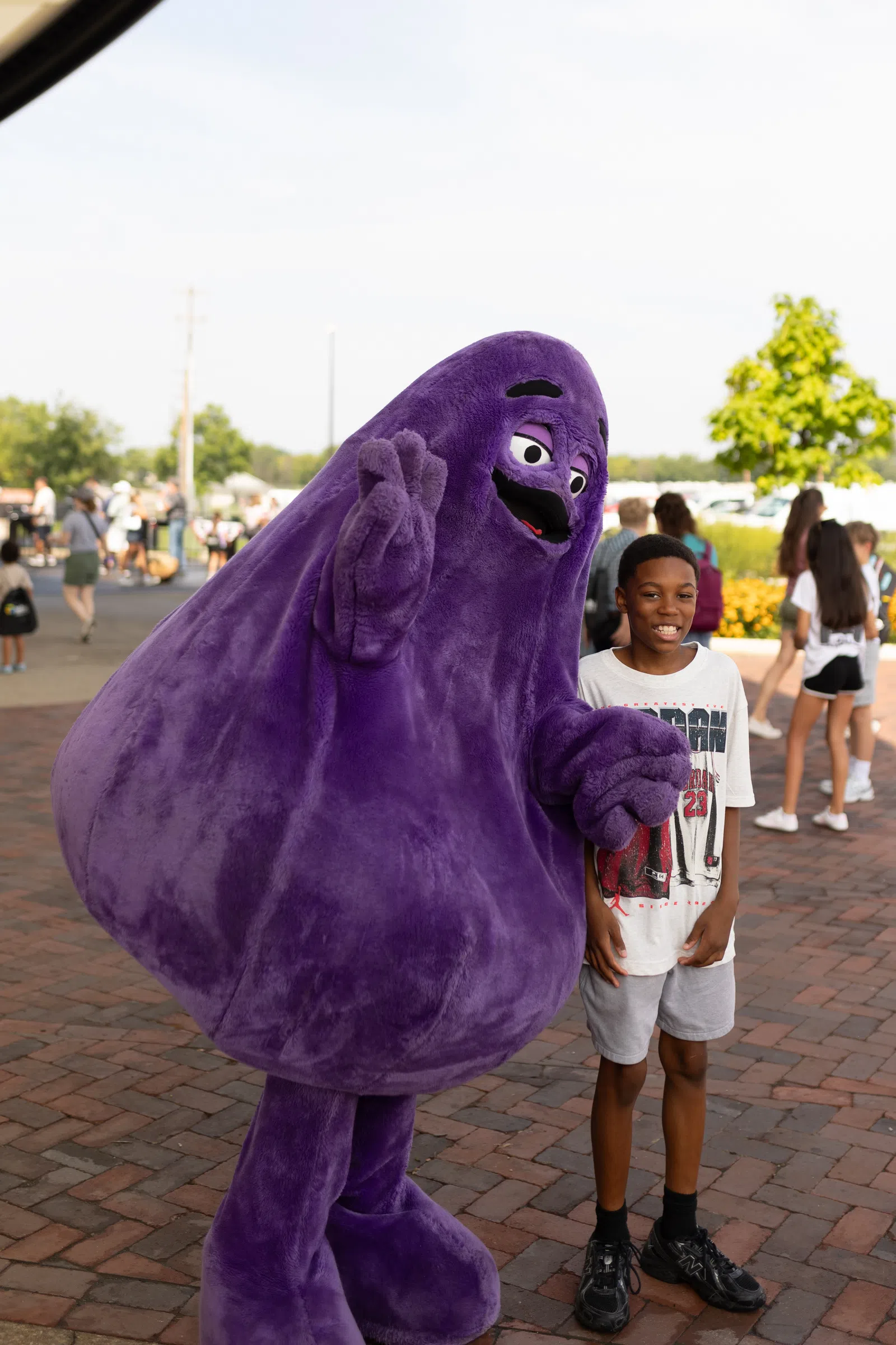 A boy posing and smiling next to a large, friendly purple mascot in an outdoor public area with people in the background.