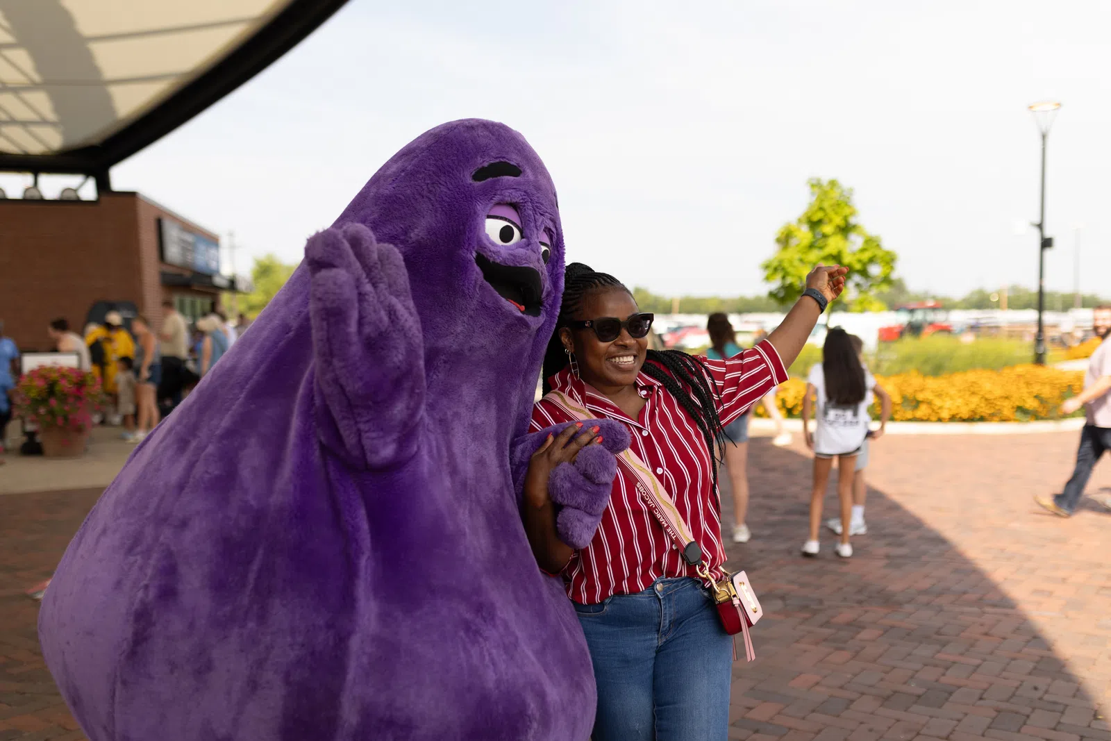 Smiling woman in sunglasses poses with a large purple mascot outdoors on a sunny day.