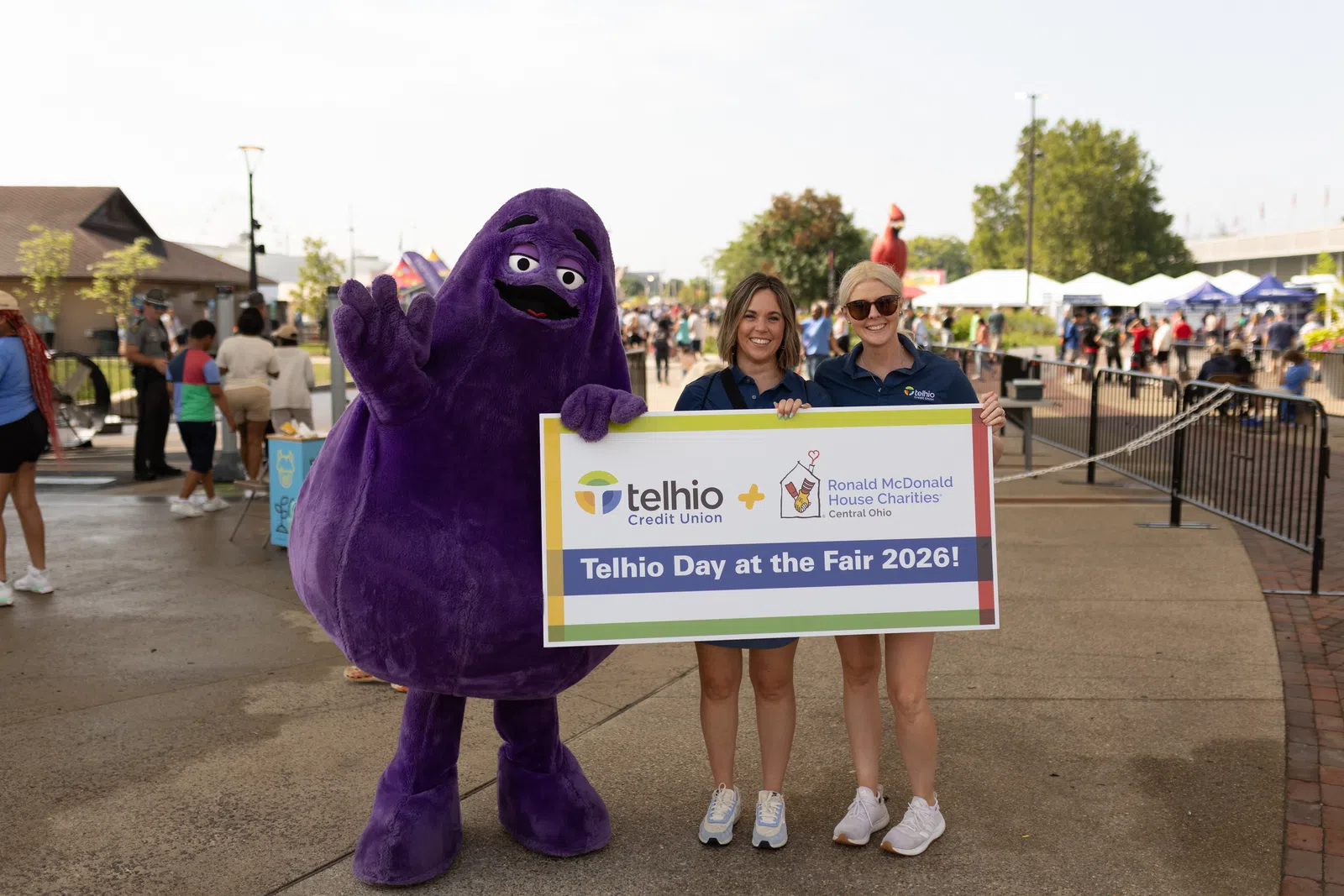 Two women and a purple mascot holding a sign promoting Telhio Day at the Fair 2026 at a crowded outdoor fair event.