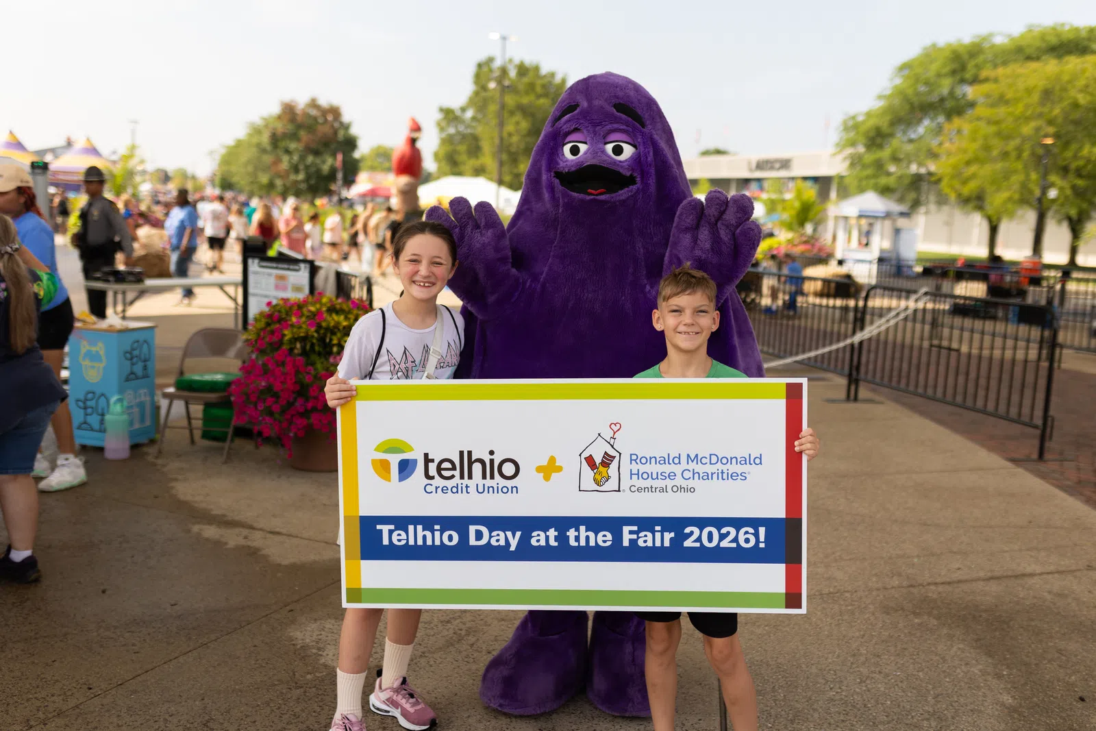 Two children and a mascot holding a Telhio Credit Union and Ronald McDonald House Charities sign at an outdoor fair.
