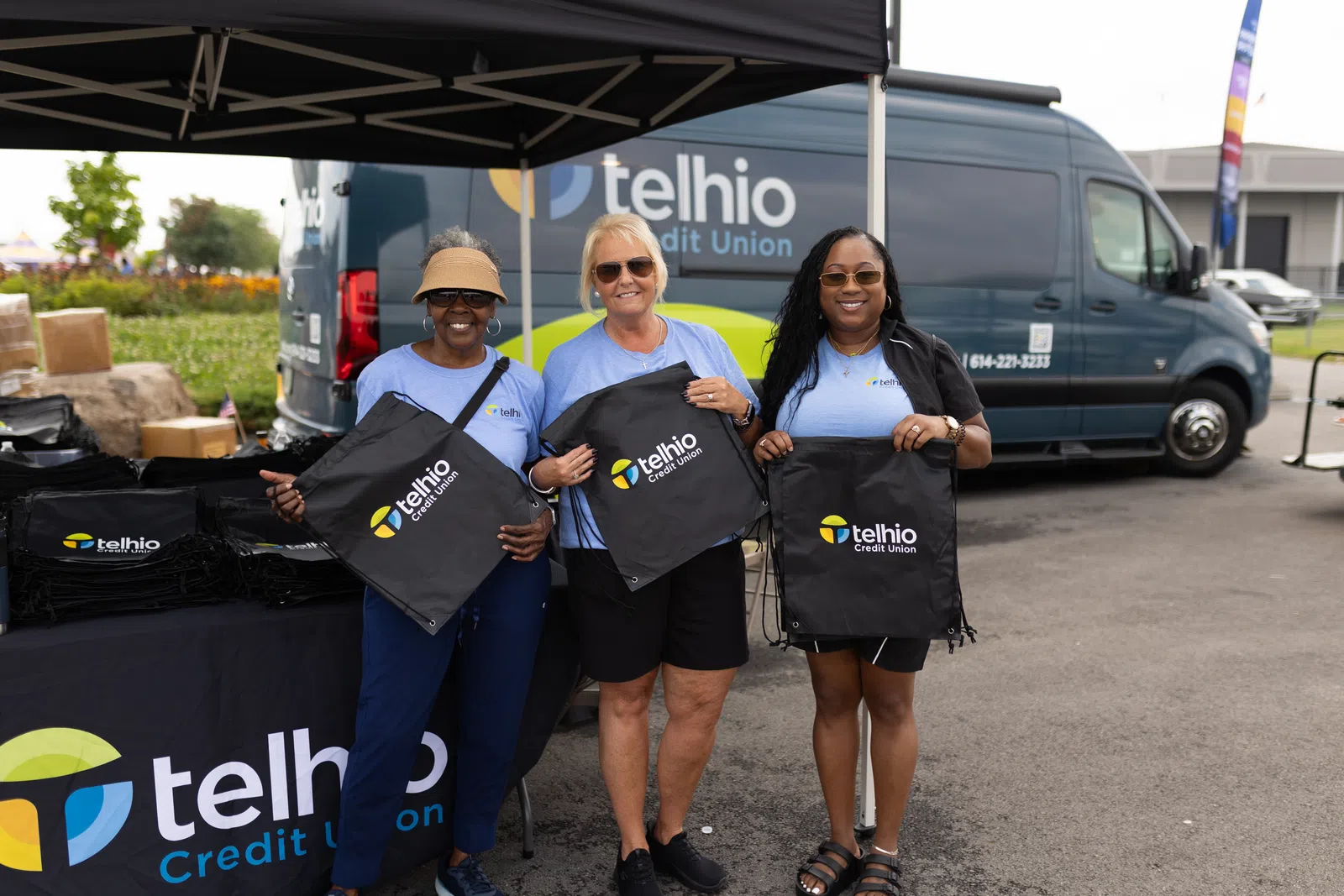 Three women holding Telhio Credit Union branded bags at an outdoor event with a Telhio van in the background